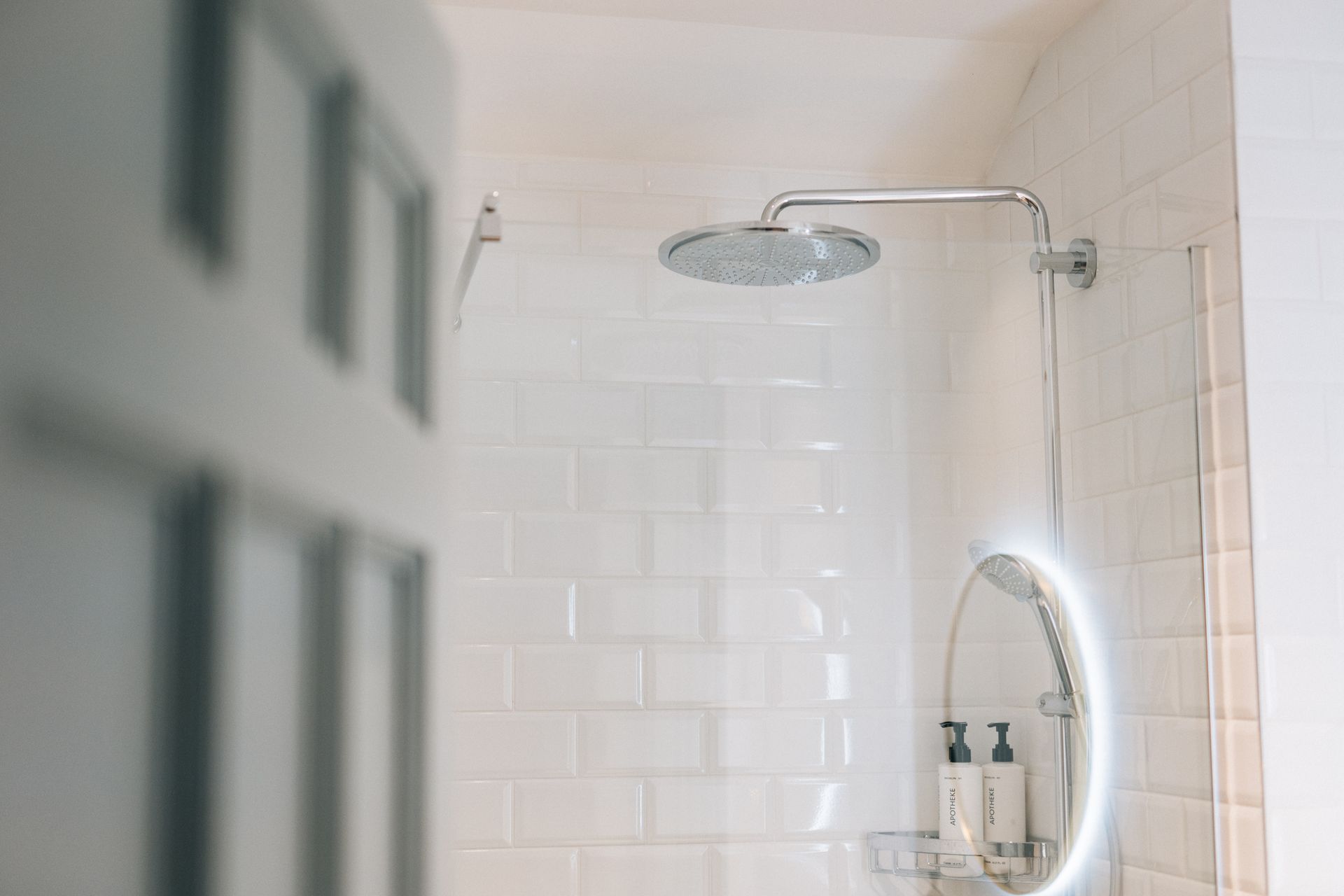 White tiled shower with rain shower head and illuminated oval mirror, seen through a partially open door.