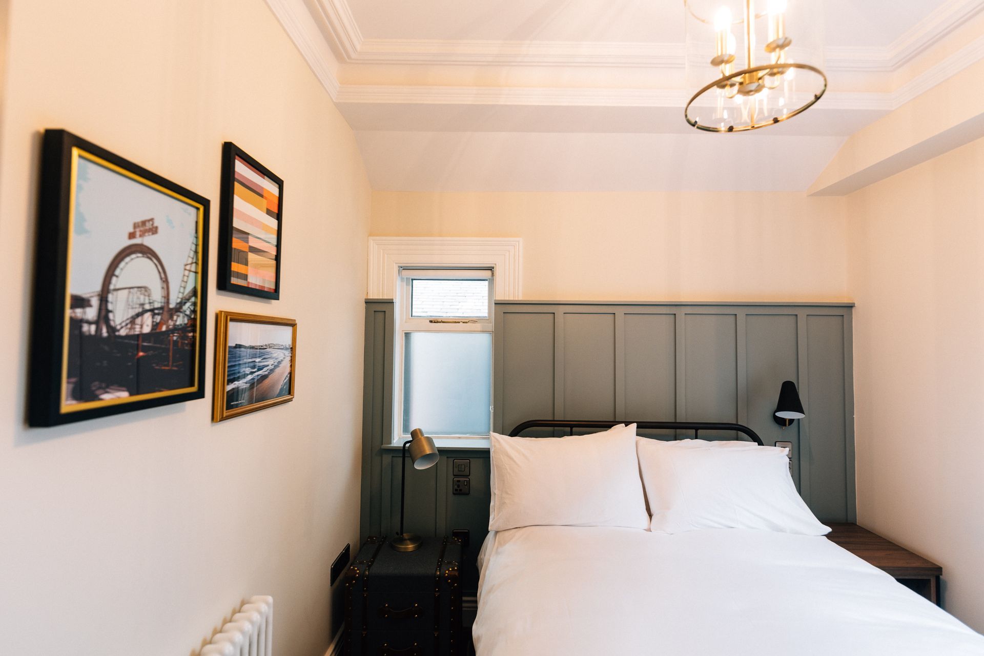 A bedroom featuring a bed with white bedding against a green paneled headboard. Artwork hangs on the cream-colored wall.