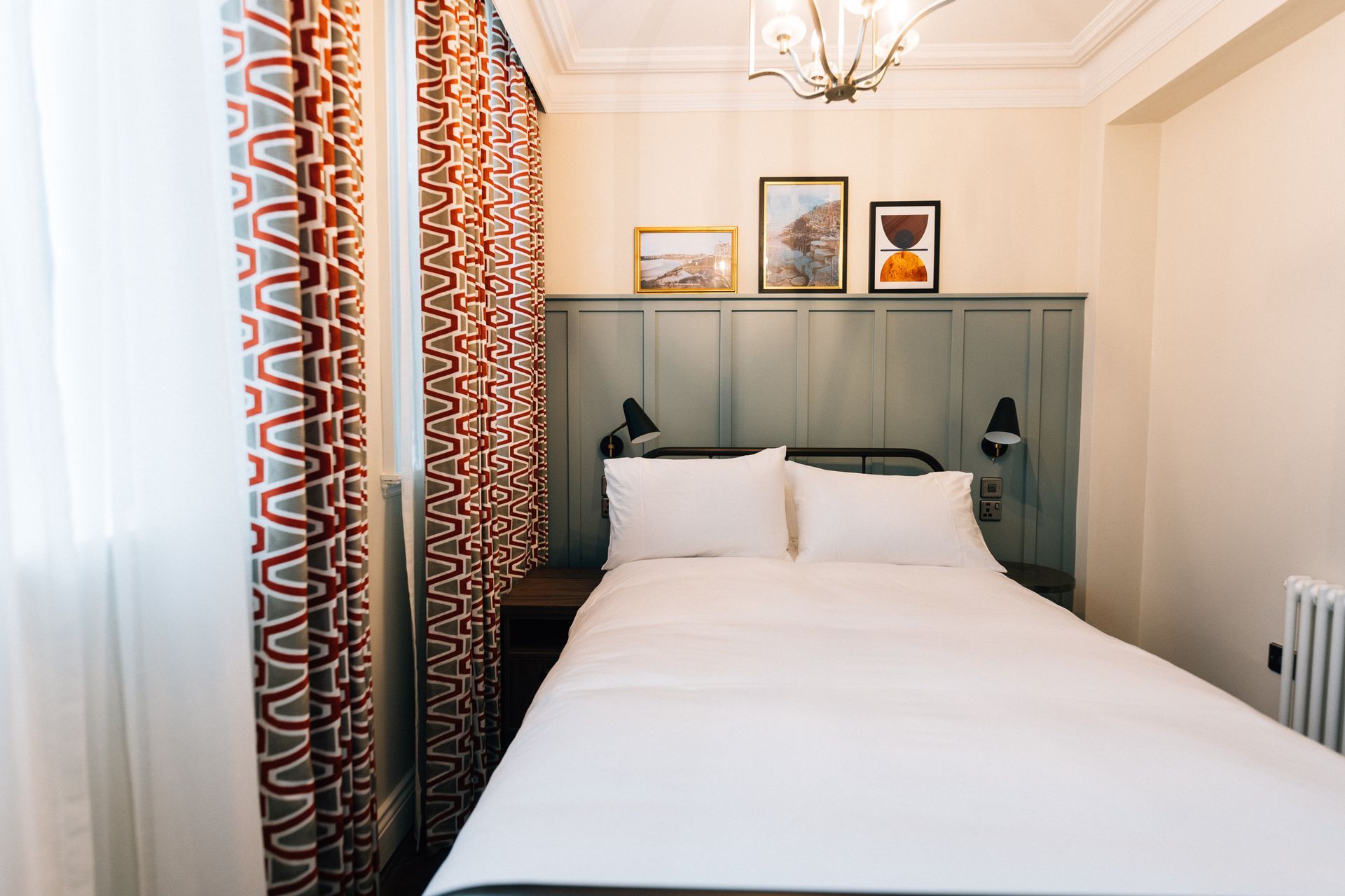Cozy hotel bedroom with a bed against a green paneled wall, framed art, patterned curtains, and a window.