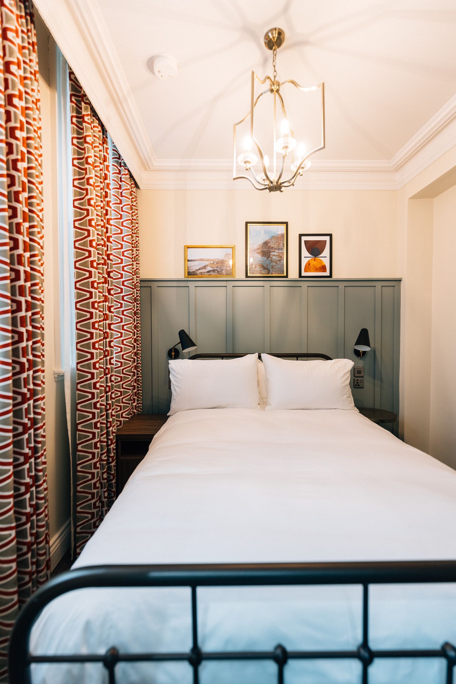 A cozy hotel bedroom with a double bed, framed pictures, patterned curtains, and a chandelier.