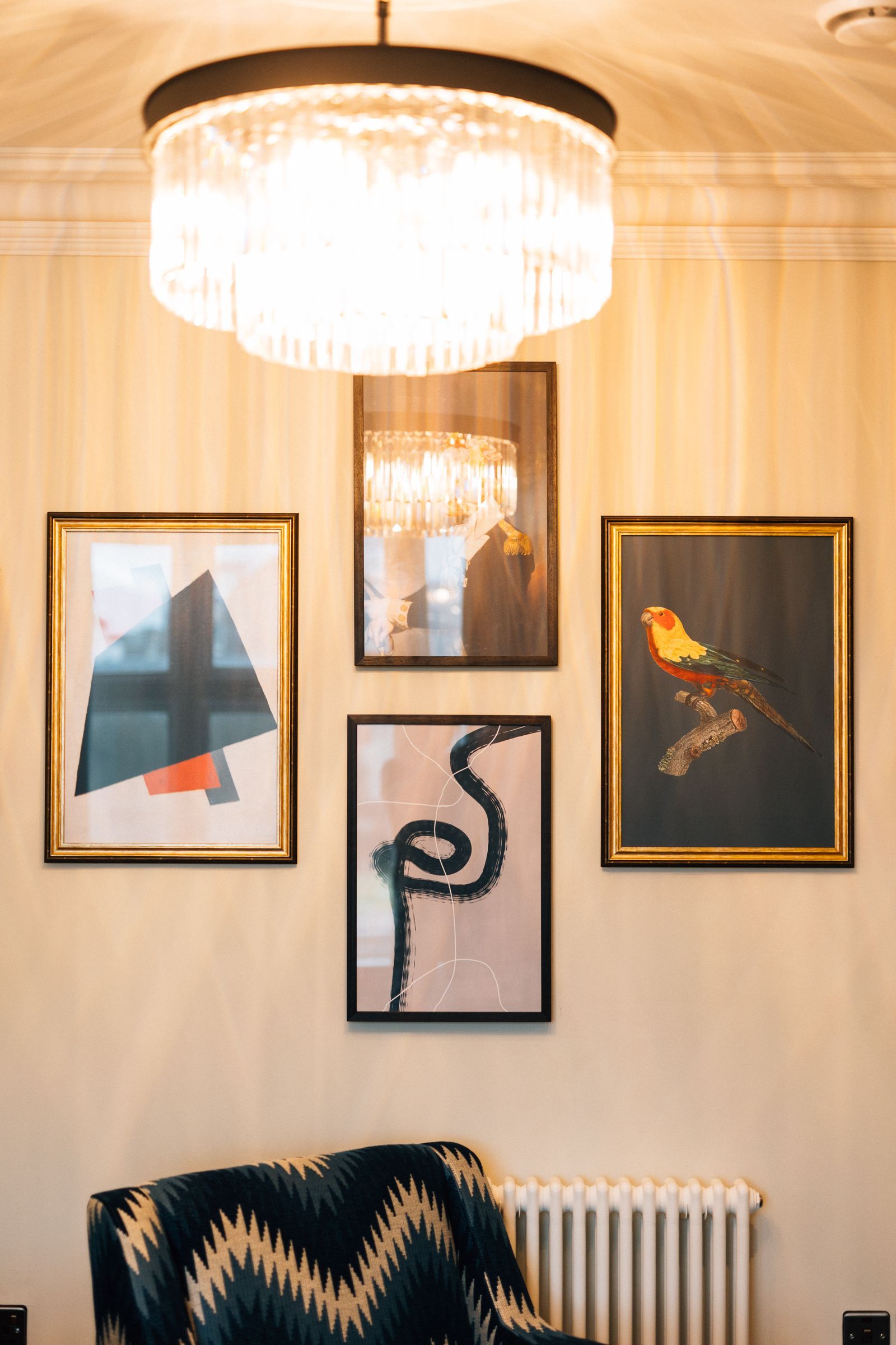 Gallery wall with framed art: abstract shapes, a bird, and a snake, lit by a round chandelier above a patterned armchair.