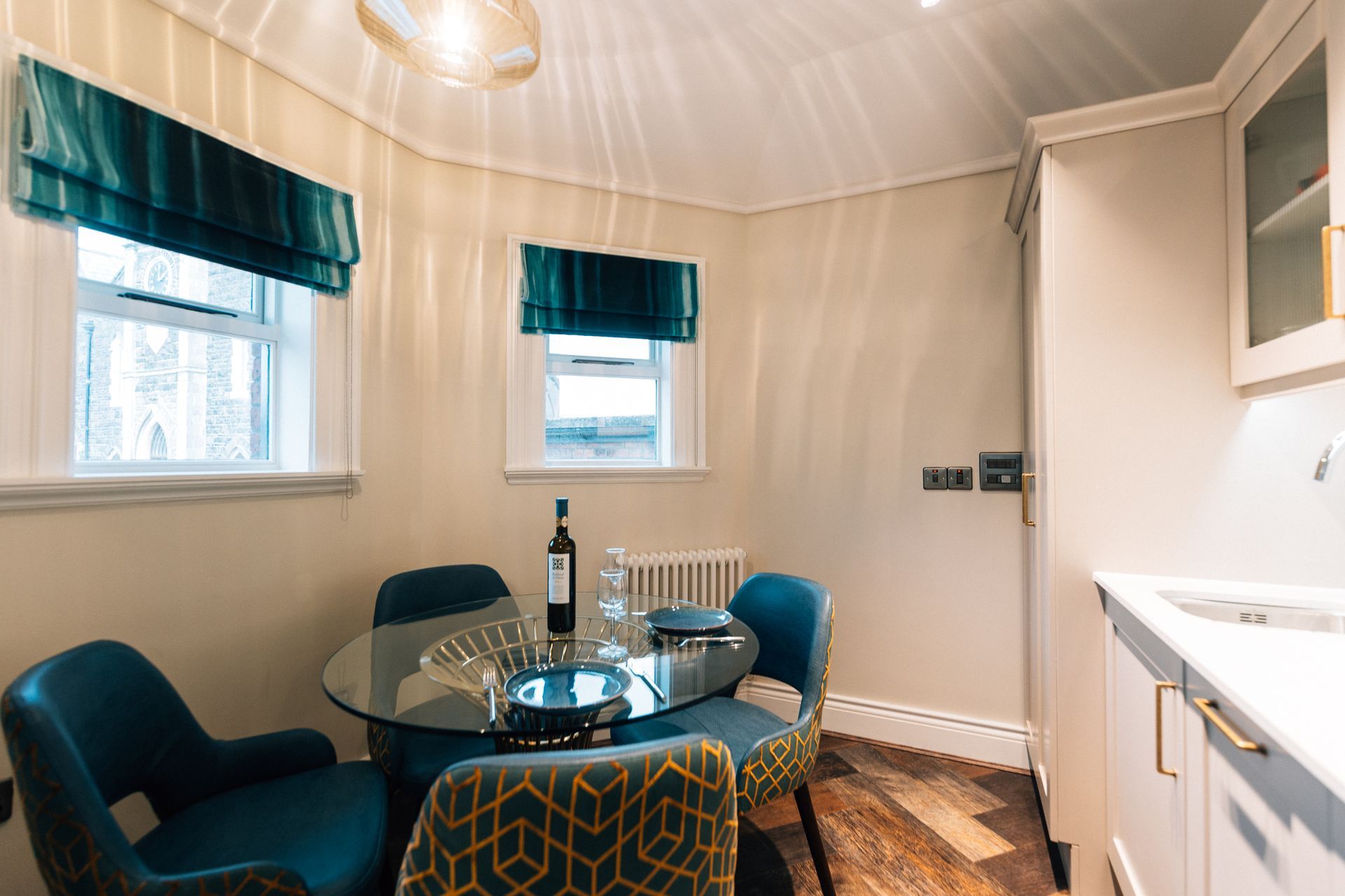 A small dining area with a glass table, four blue chairs, and two windows with teal shades. 