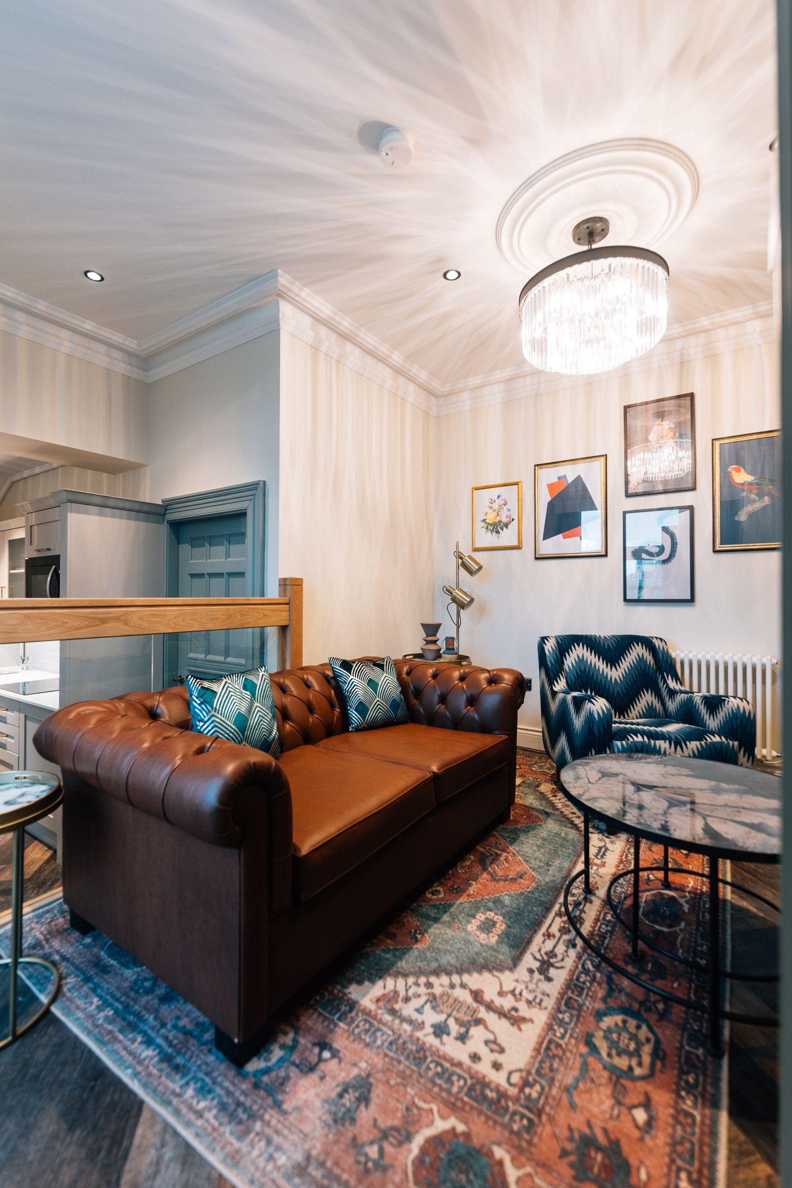 Living room with a brown leather Chesterfield sofa, patterned rug, artwork, and a crystal chandelier.