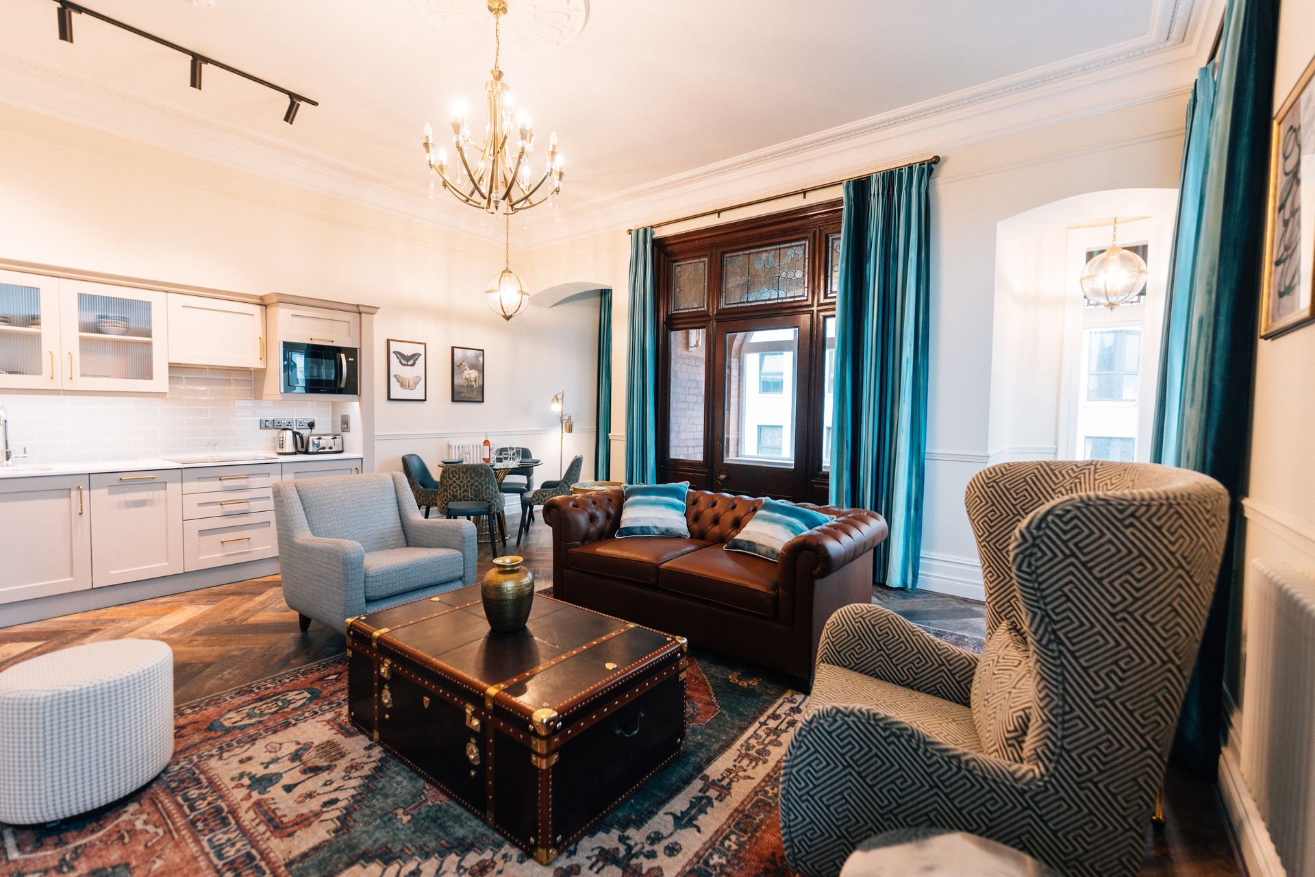 Elegant living room with blue chairs, brown sofa, ornate rug, and large windows with teal curtains