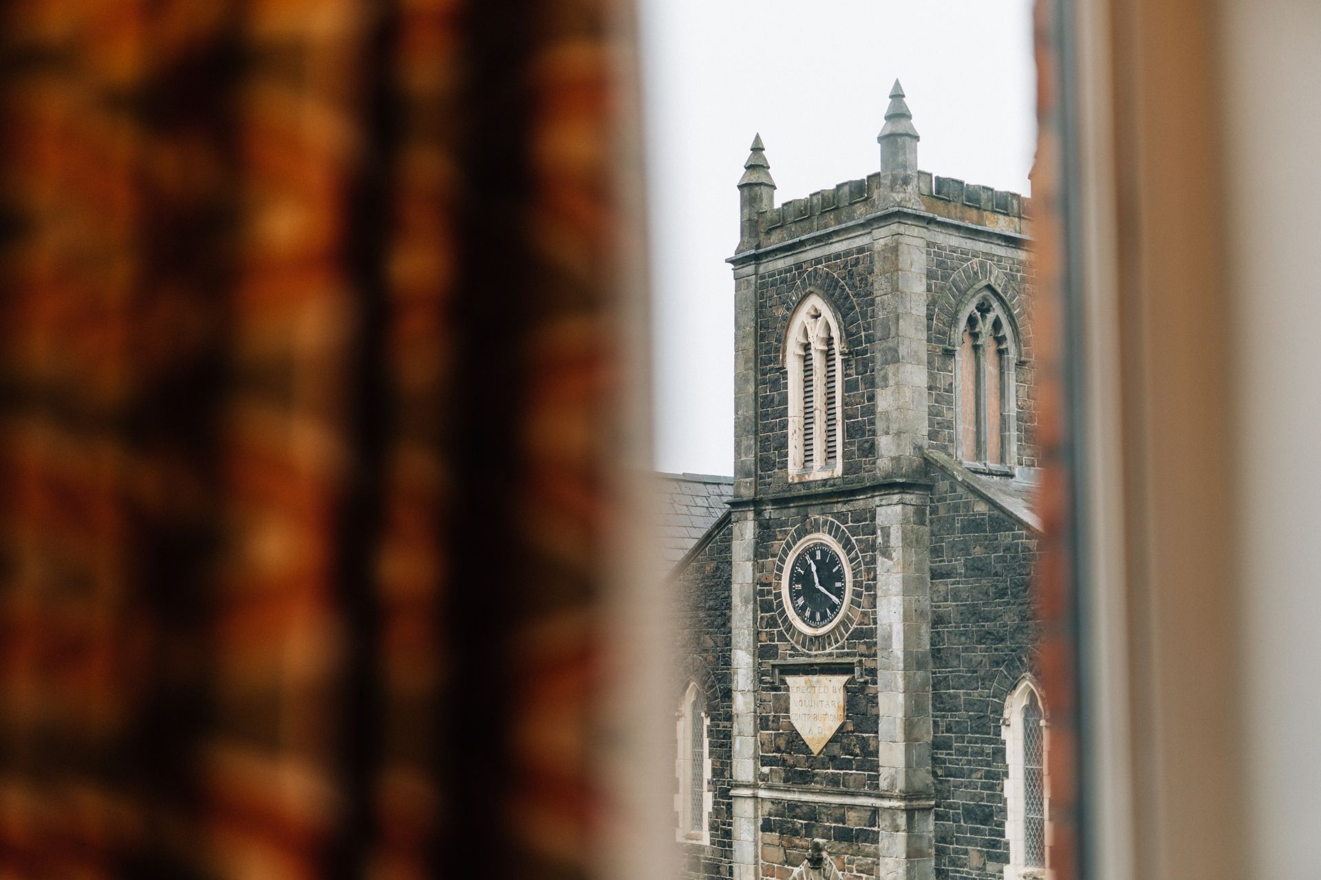 View of a stone church tower with a clock through a window, framed by patterned curtains.