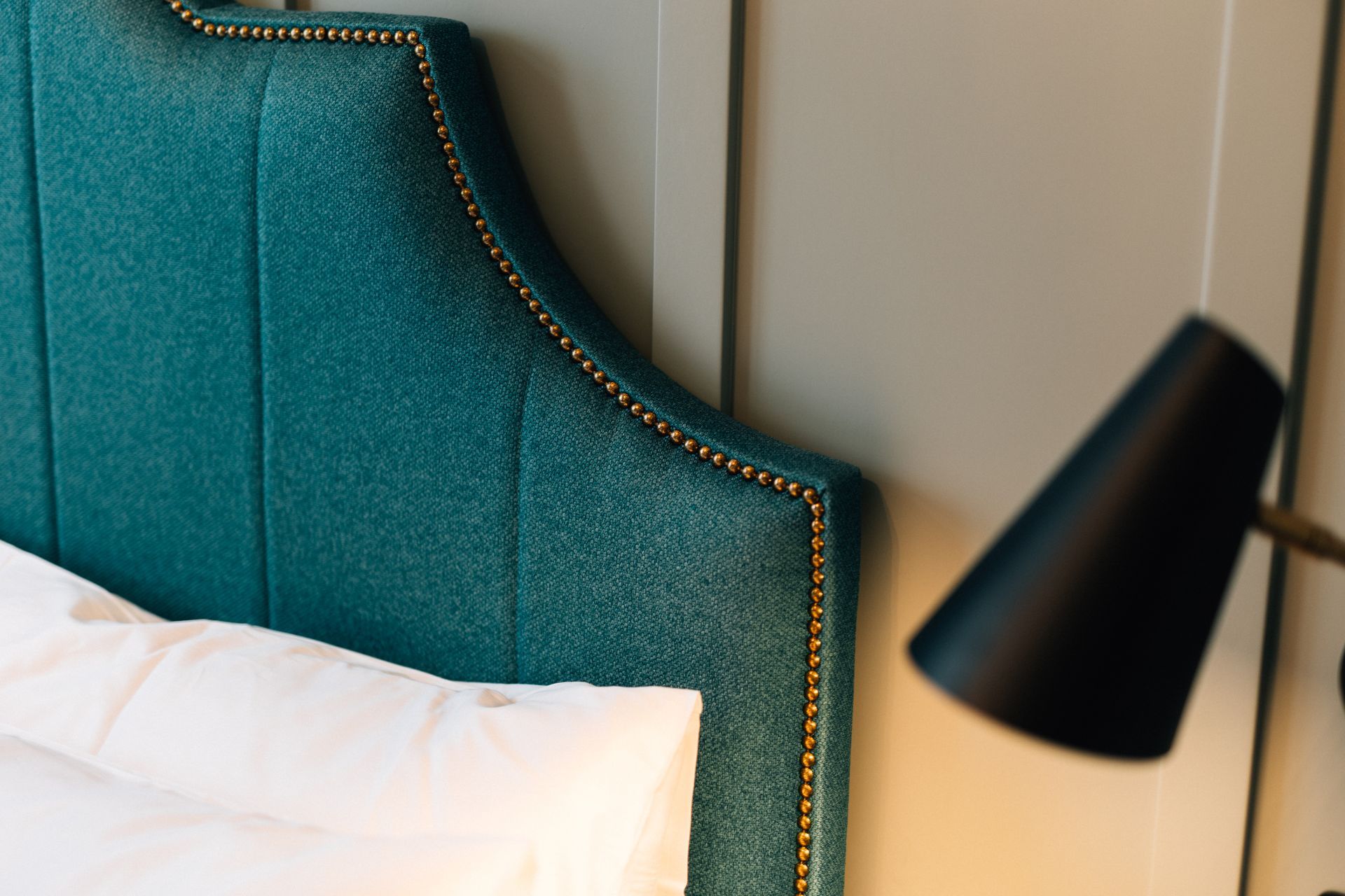 Teal upholstered headboard with gold nailhead trim; a white pillow rests on the bed with a black lamp in the background.