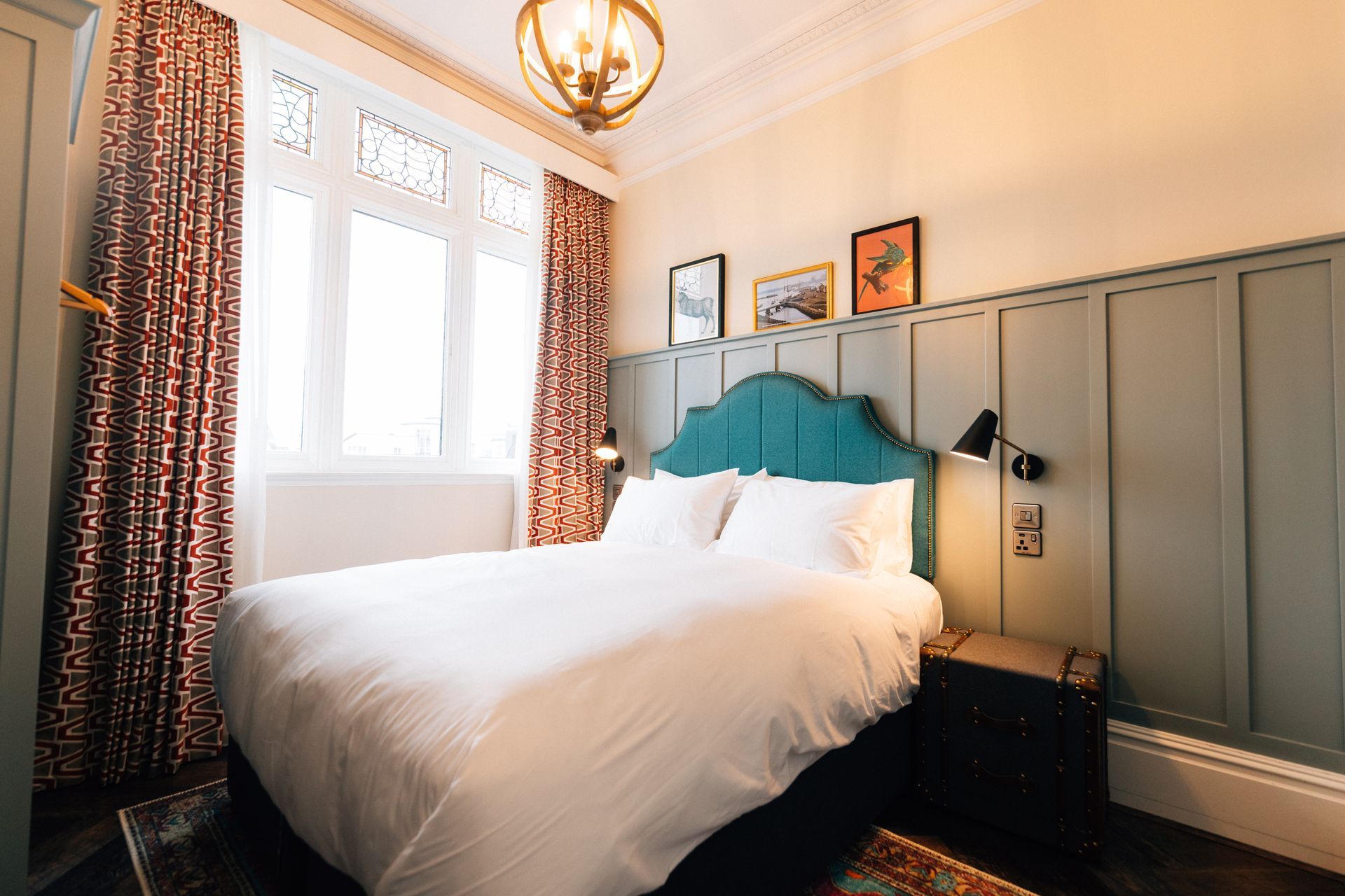 A cozy bedroom with a bed against a green panel wall. A teal headboard is centered under framed artwork. 
