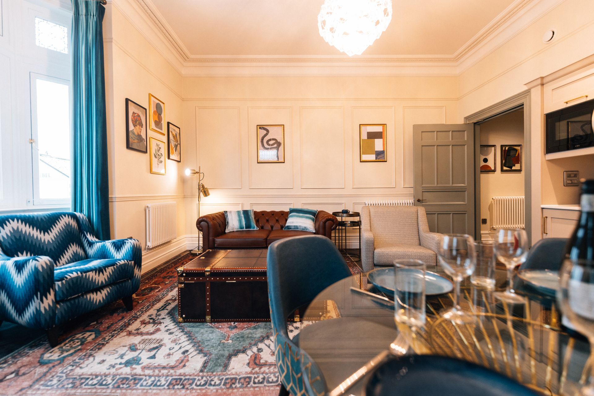 A living room with a patterned blue sofa, leather trunk, and dining table; decorated with artwork and a chandelier.