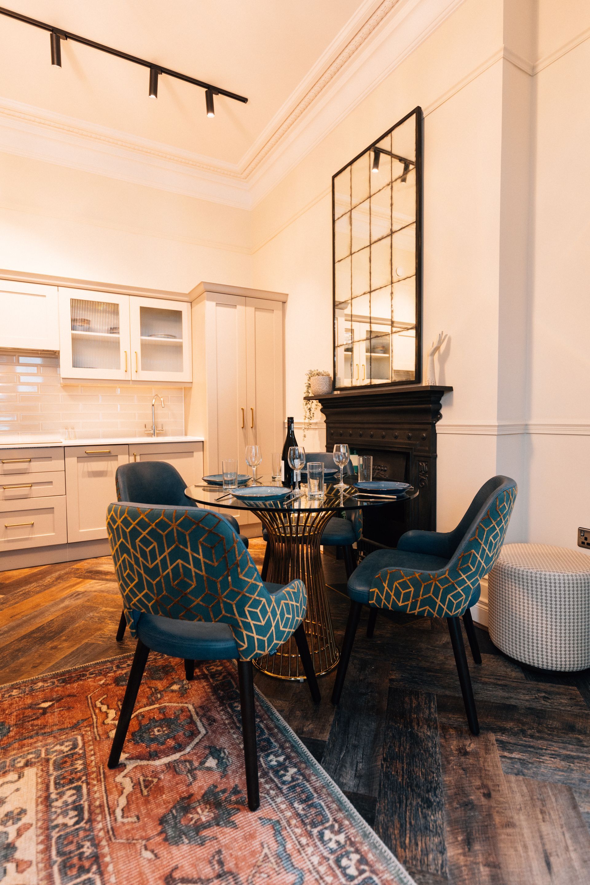 A dining area with a table and patterned chairs on a rug, kitchen in the background, large mirror on the wall.