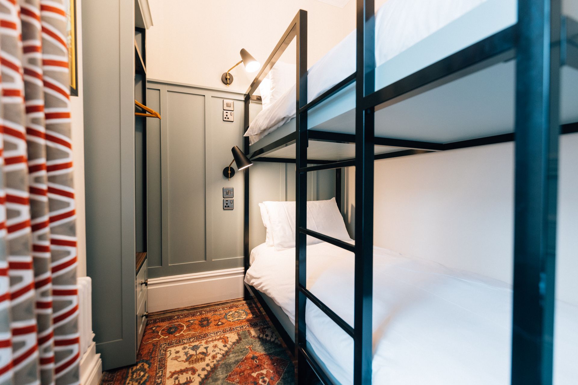 Narrow hostel room with bunk beds, colorful rug, and wall paneling. Curtains with a red and white pattern are visible.