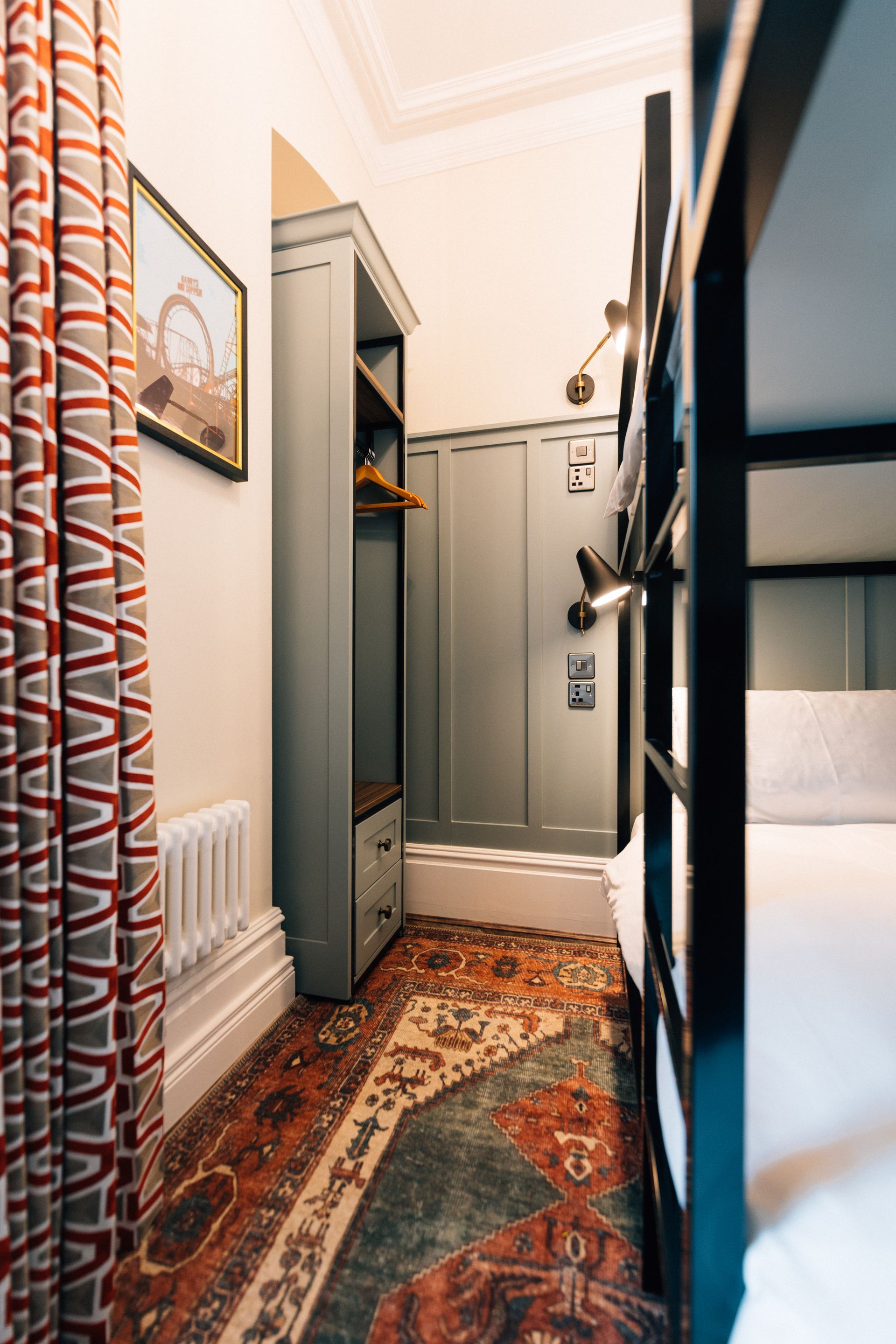 Narrow hostel room with bunk beds, a patterned rug, and a built-in wardrobe; a window with patterned curtains is visible.