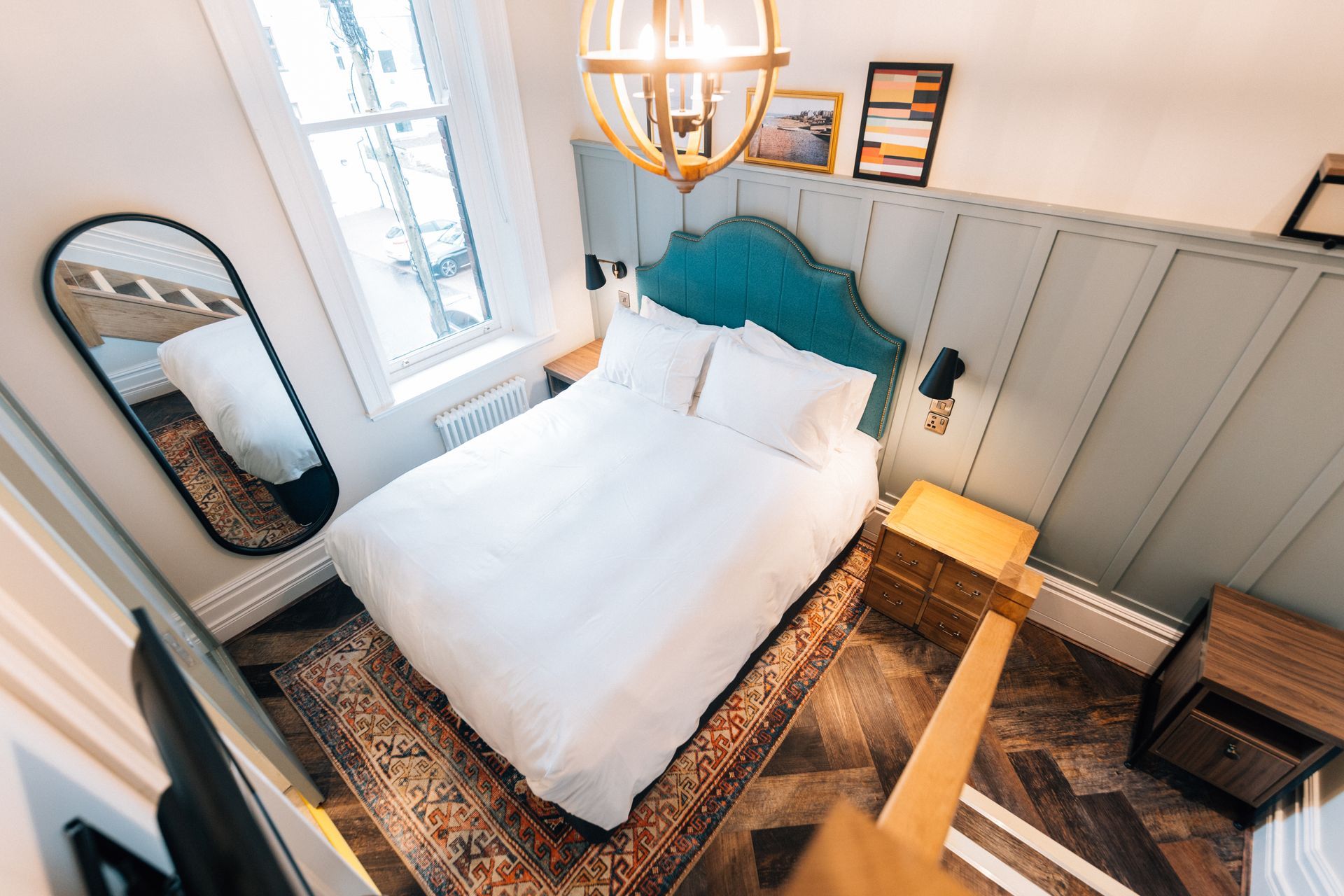 Cozy bedroom with a bed, mirror, and bedside tables; a window to the left; overhead light fixture.