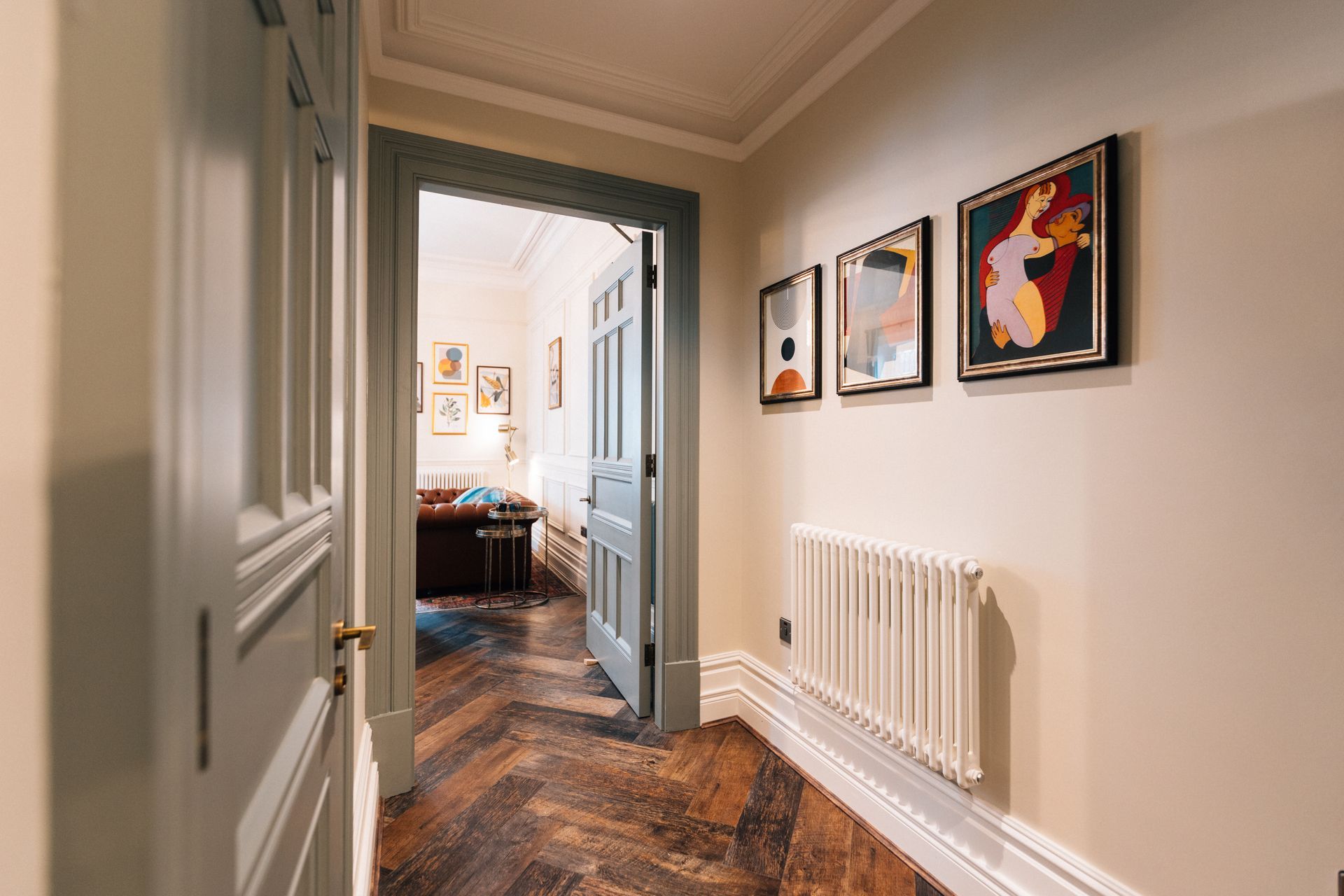 Hallway with herringbone wood floor, light-colored walls, and framed art. An open door leads to another room.