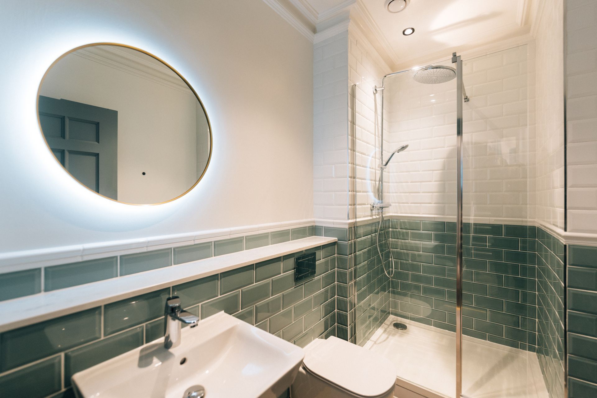 A modern bathroom featuring a circular mirror with a glowing rim, a shower, a sink, and gray tiled walls.