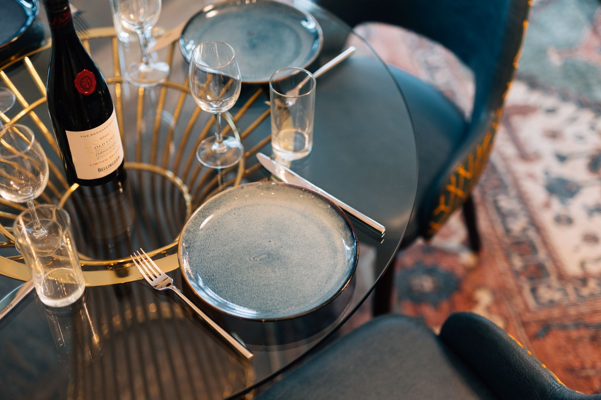 A round glass dining table set with plates, silverware, glasses, and a bottle of wine. The setting is indoors.