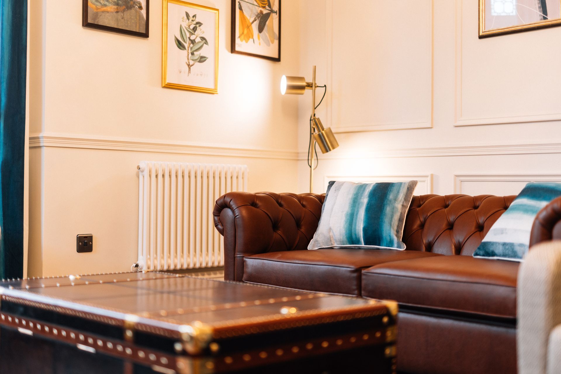 A luxurious living room with a brown leather Chesterfield sofa, art, and a brass floor lamp. 