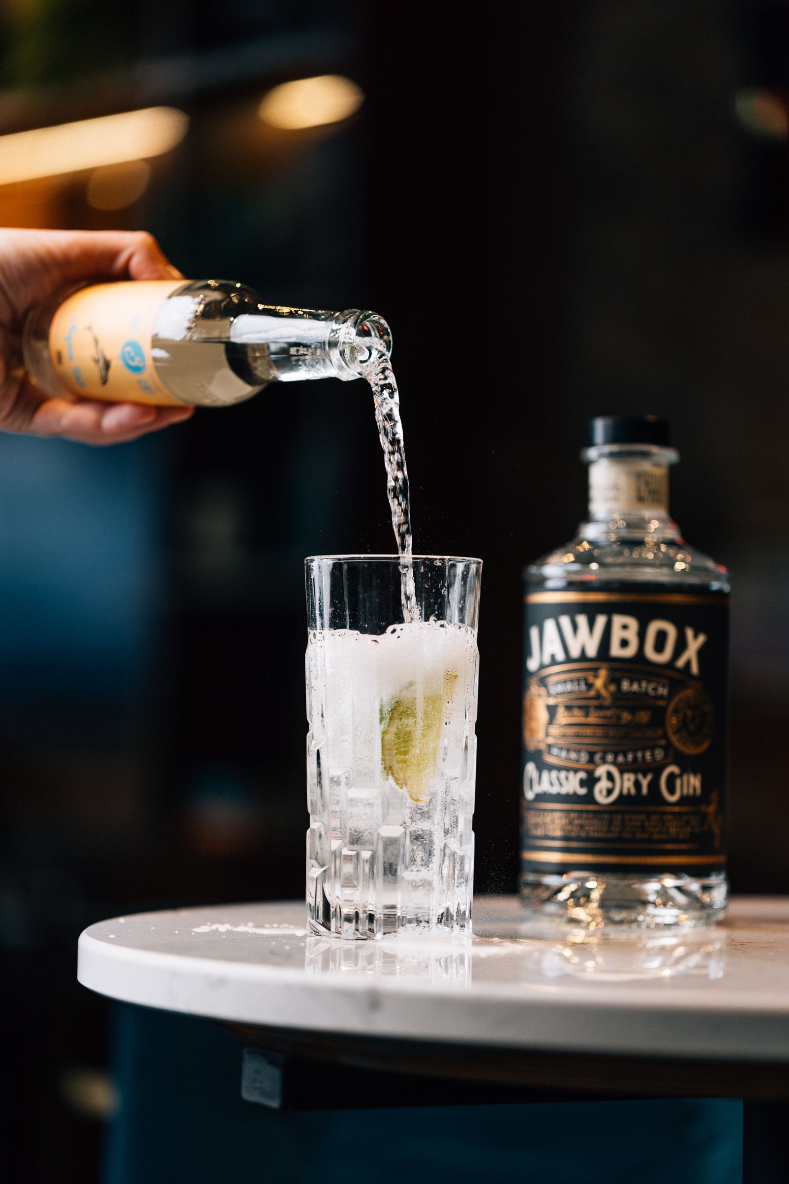 A hand pouring clear liquid from a small bottle into a glass with ice and lime. 