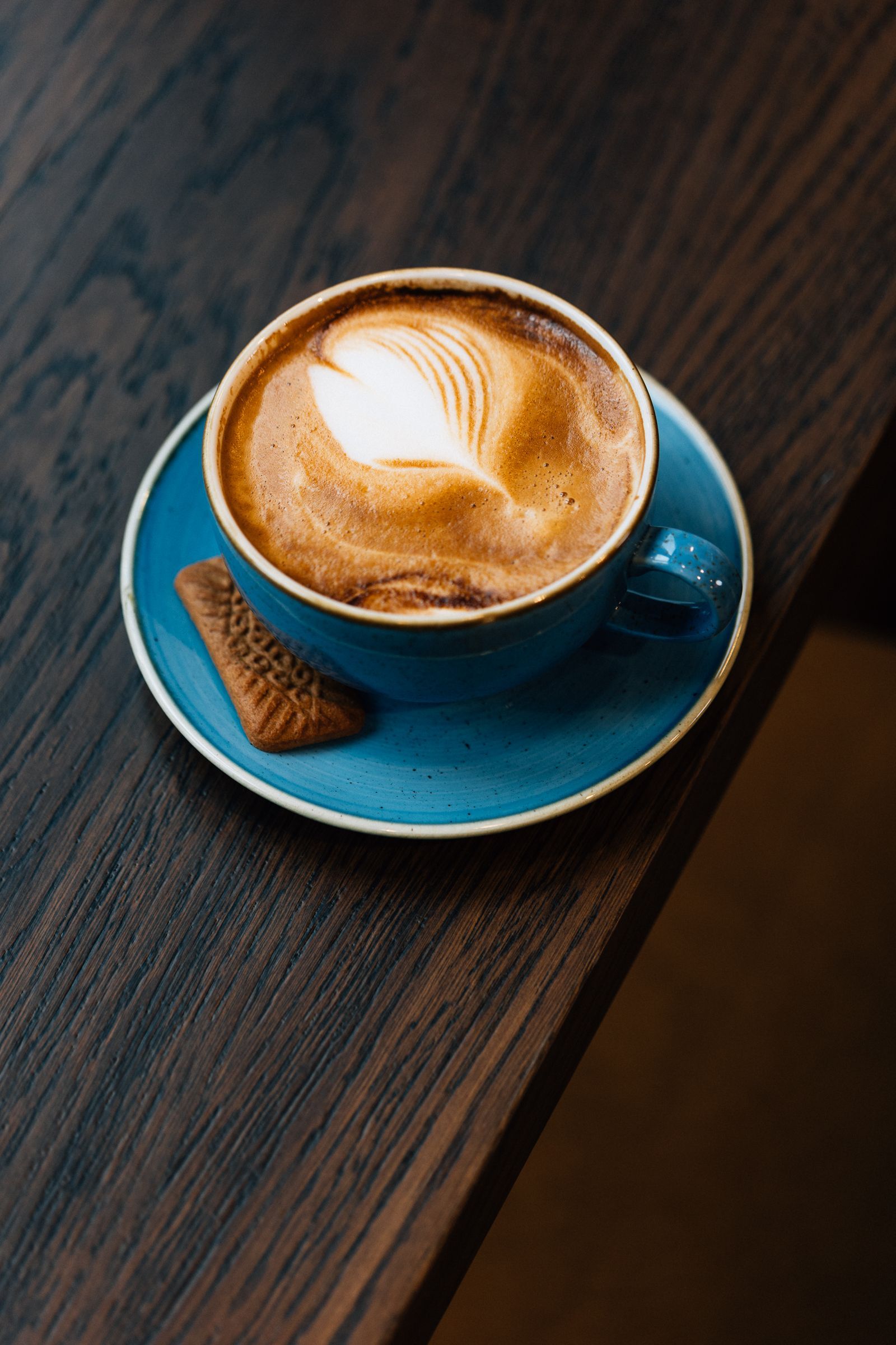 A cup of cappuccino with latte art and a cookie, on a blue saucer, resting on a dark wooden table.