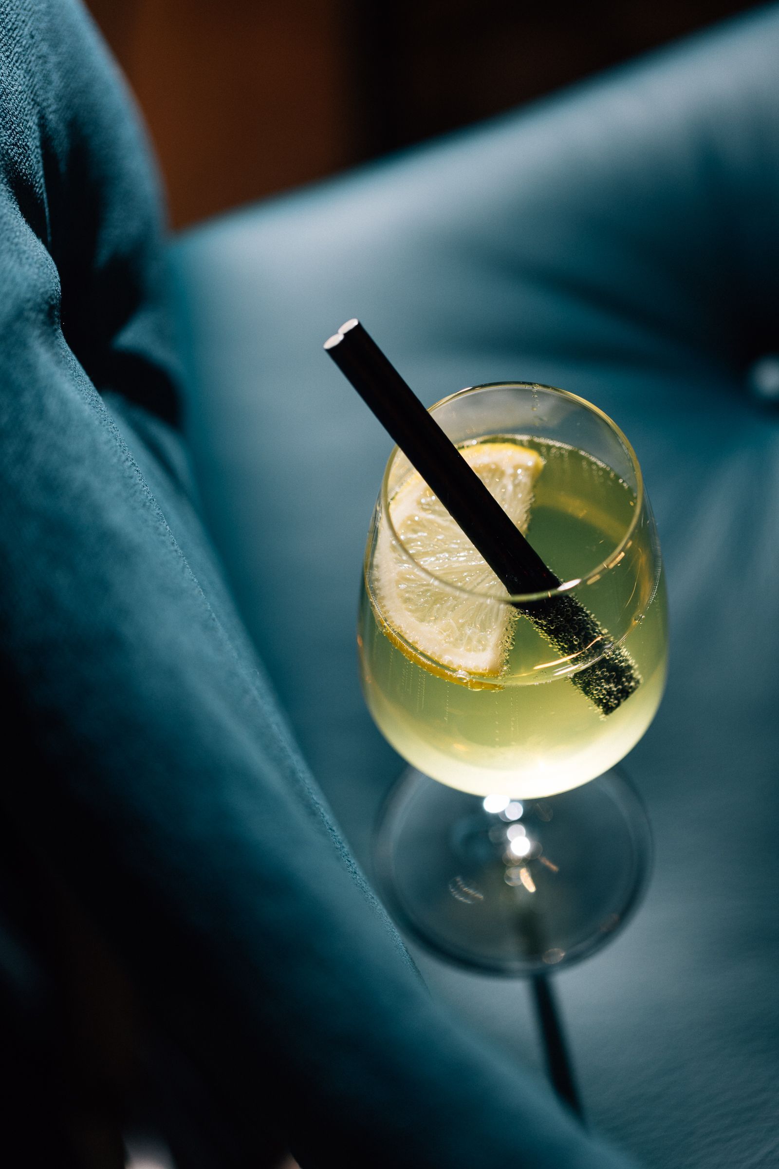 A cocktail in a glass with a lemon slice and black straw, placed on a reflective surface.