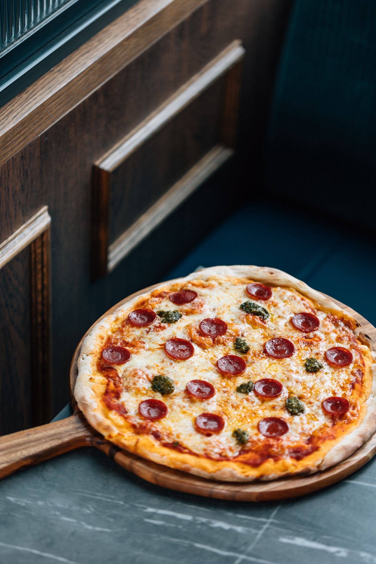 Pepperoni pizza on a wooden serving board, resting on a marble table with blue booth seating in the background.