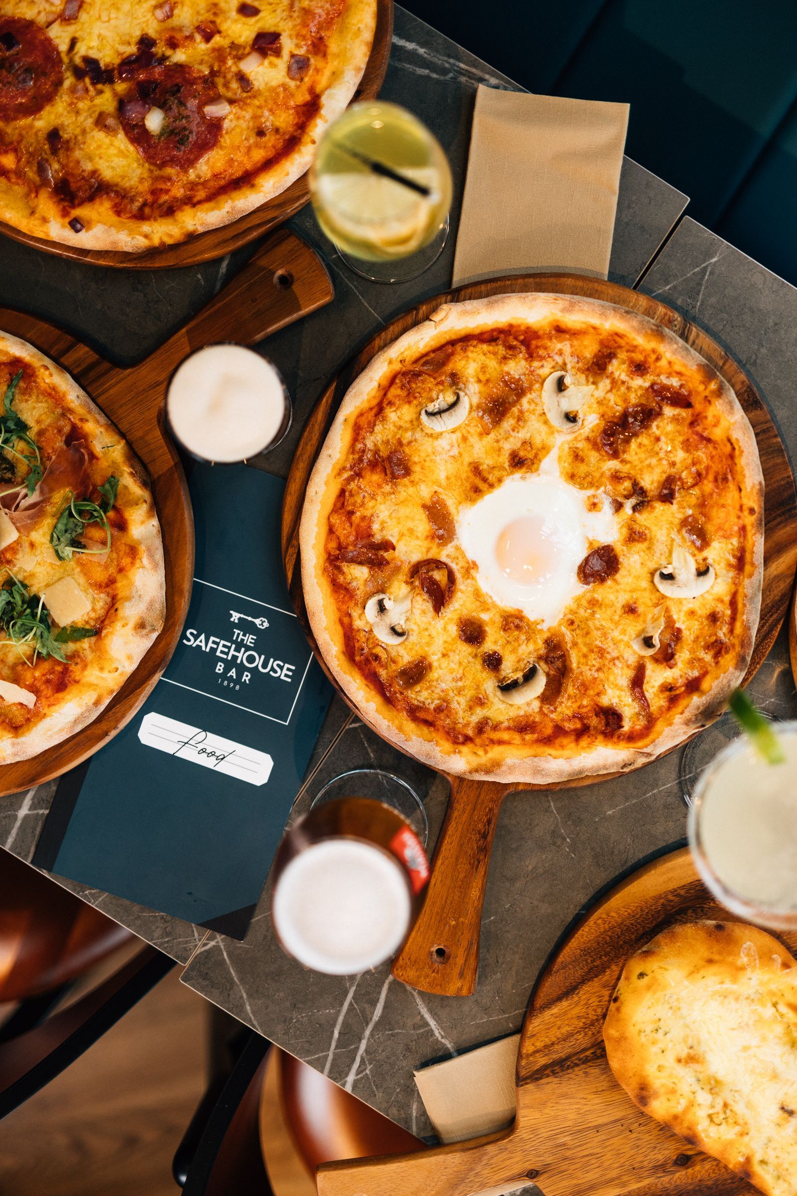 Overhead view of multiple pizzas on wooden boards with drinks on a checkered table.
