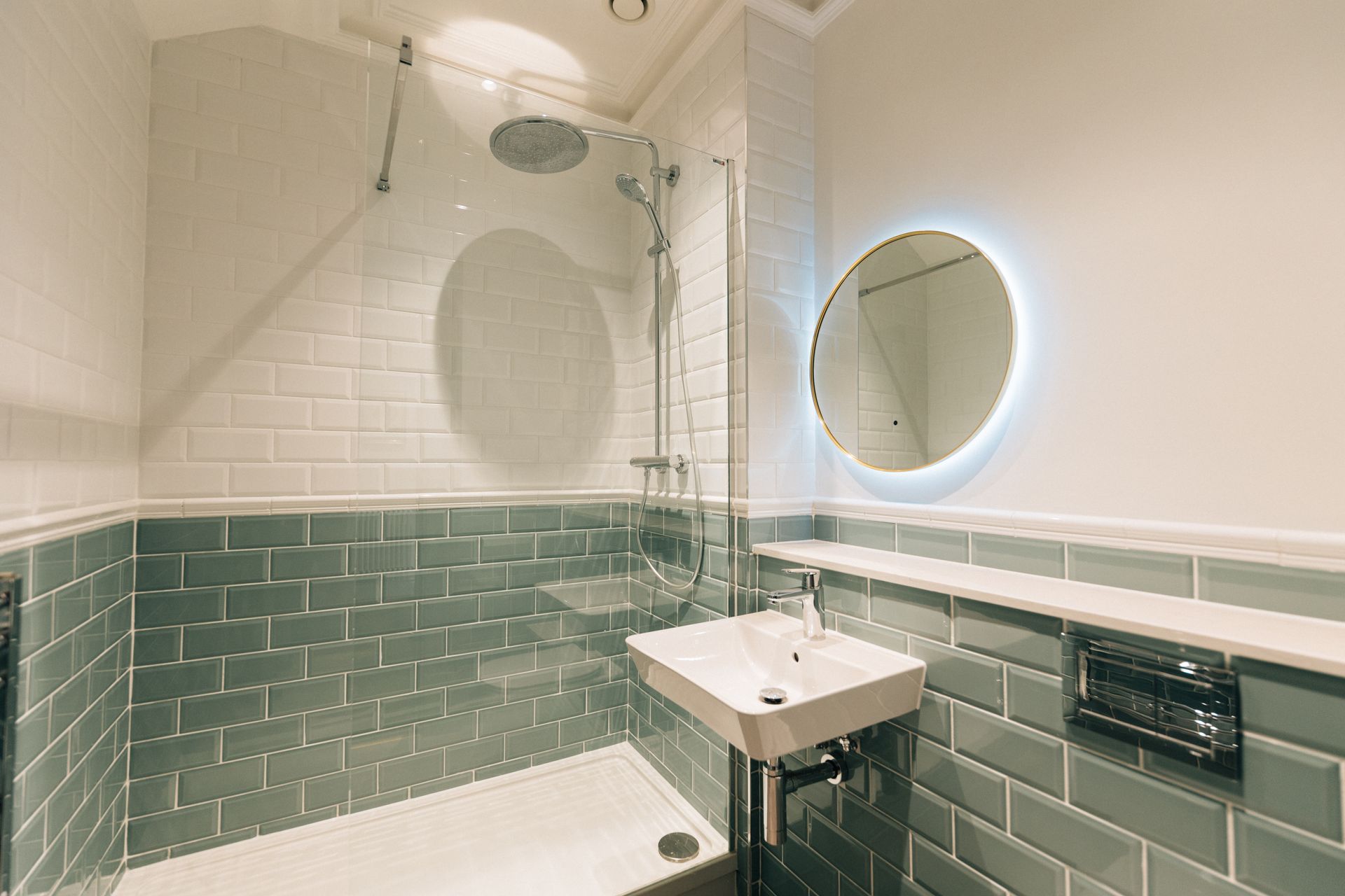 Modern bathroom with green tile, white tub, glass shower, and round illuminated mirror by a wall sink