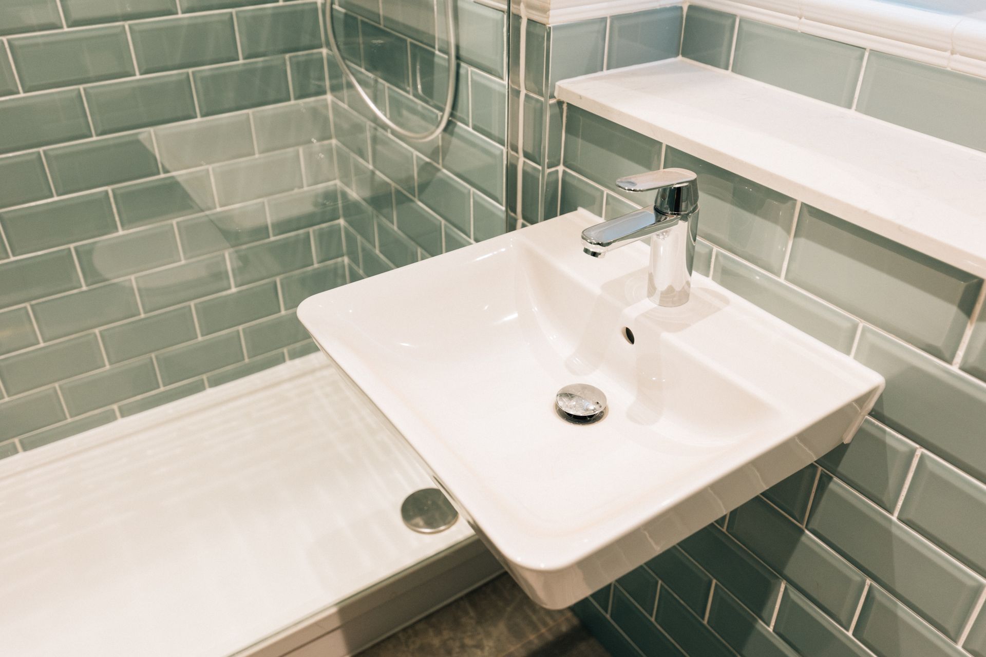 A small, modern bathroom with a white sink and a glass-enclosed shower, light green tiled walls, and a white shelf.