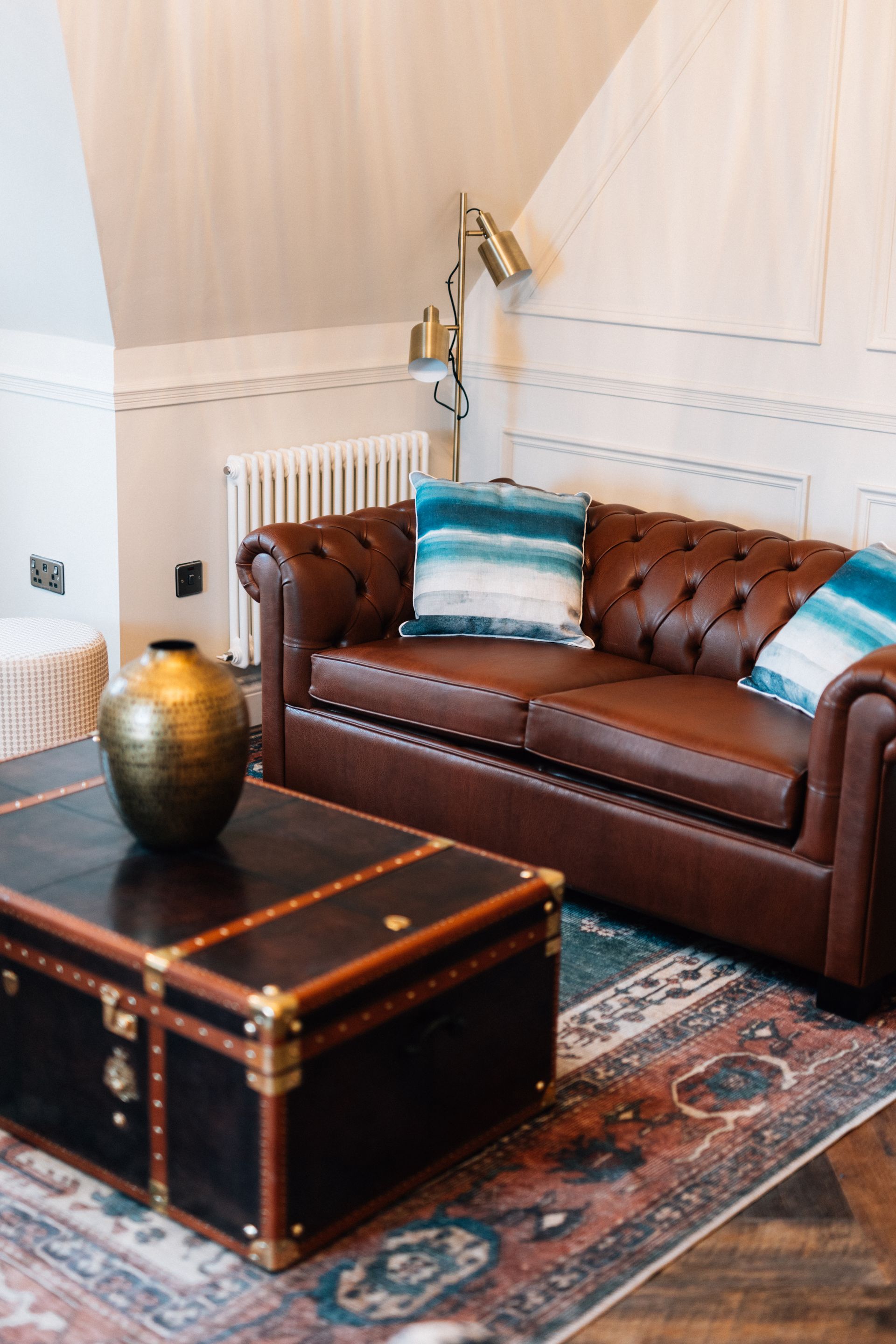 A brown leather Chesterfield sofa with two blue-toned pillows sits in a room with a trunk coffee table and a patterned rug.