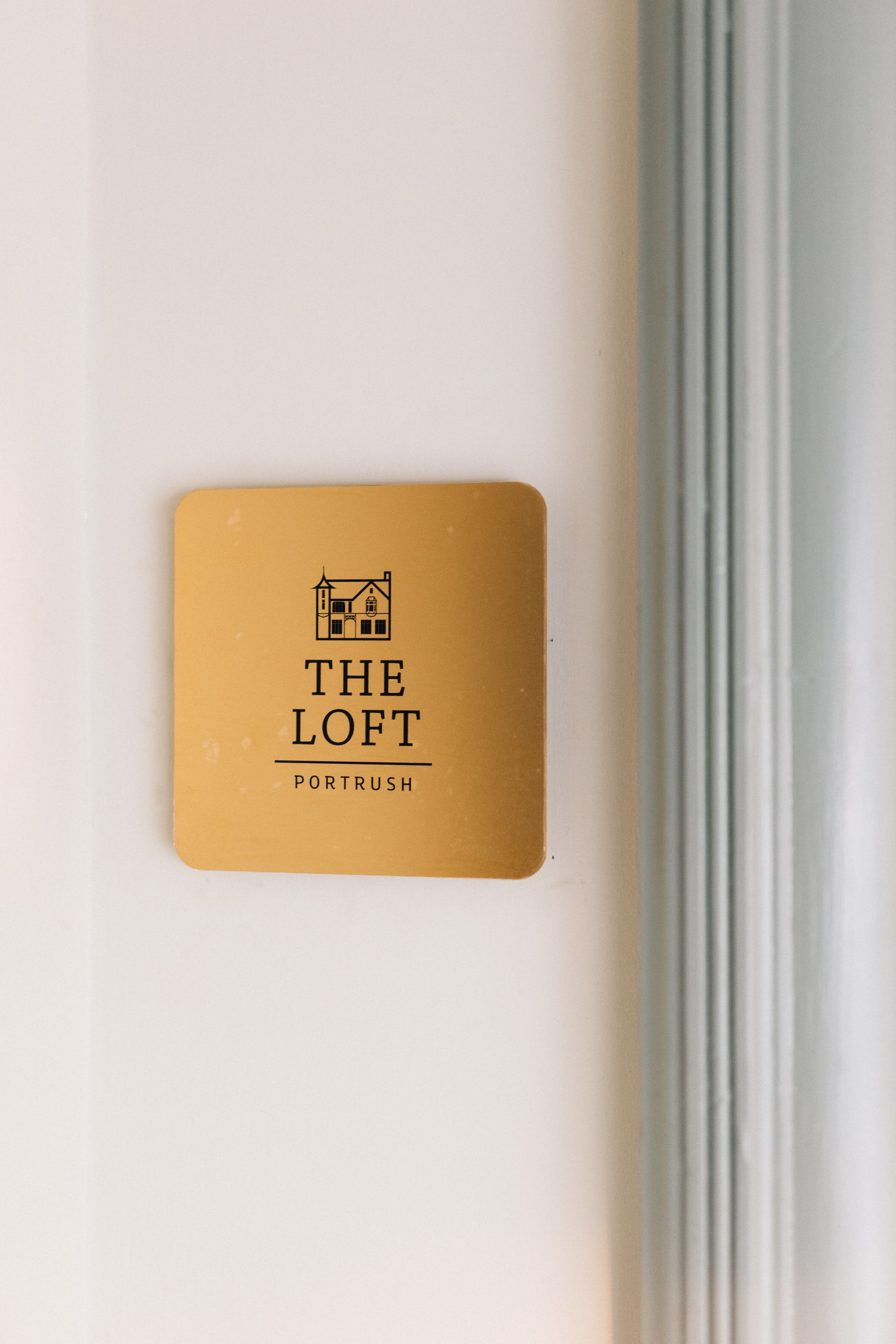 Gold square sign reading “THE LOFT” on a white wall beside a gray doorframe