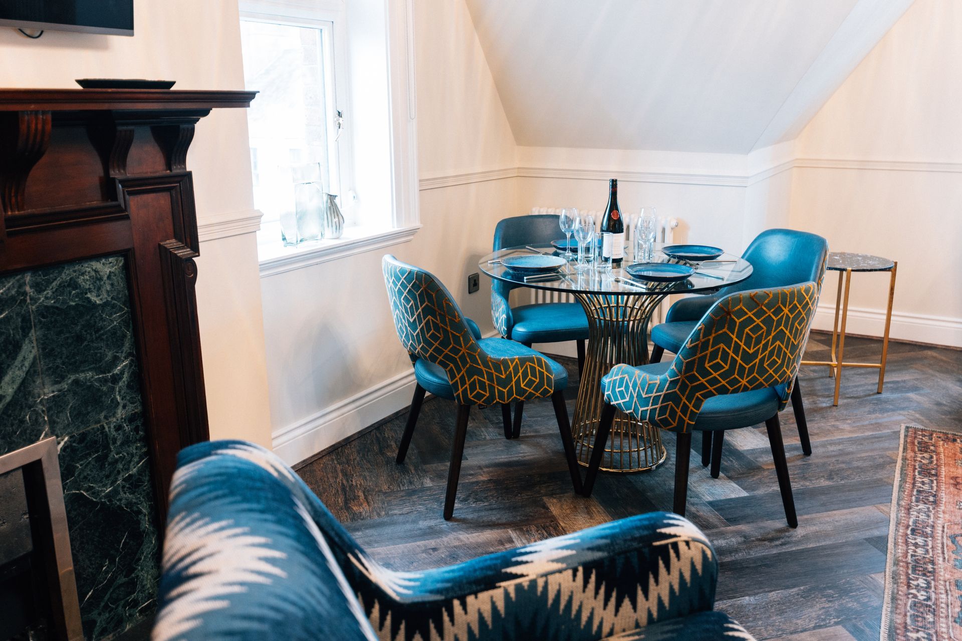 Cozy dining area with a round glass table set for four, teal patterned chairs, and a dark wooden fireplace.