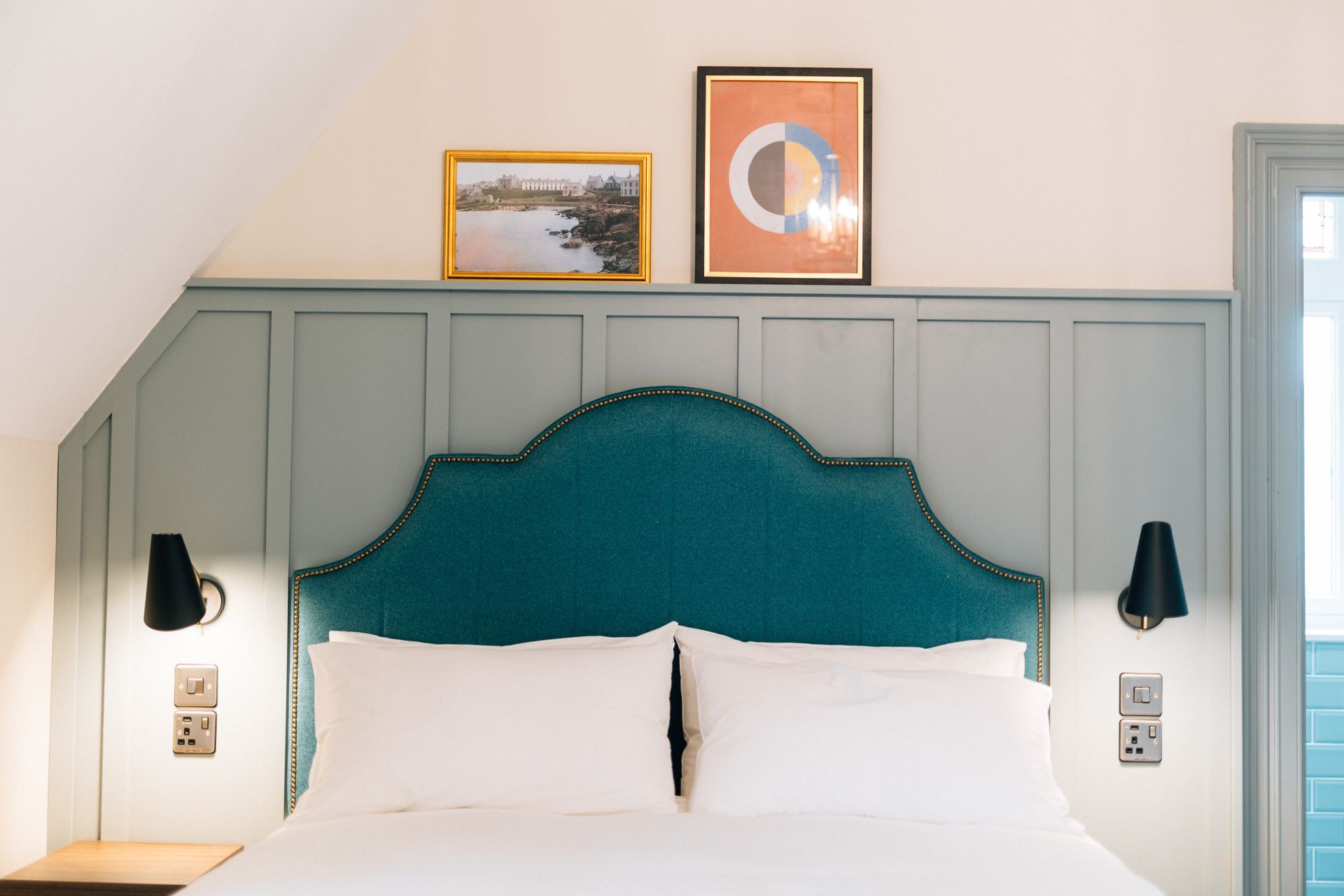 A cozy bedroom with a teal headboard, white bedding, and two wall-mounted lamps. 