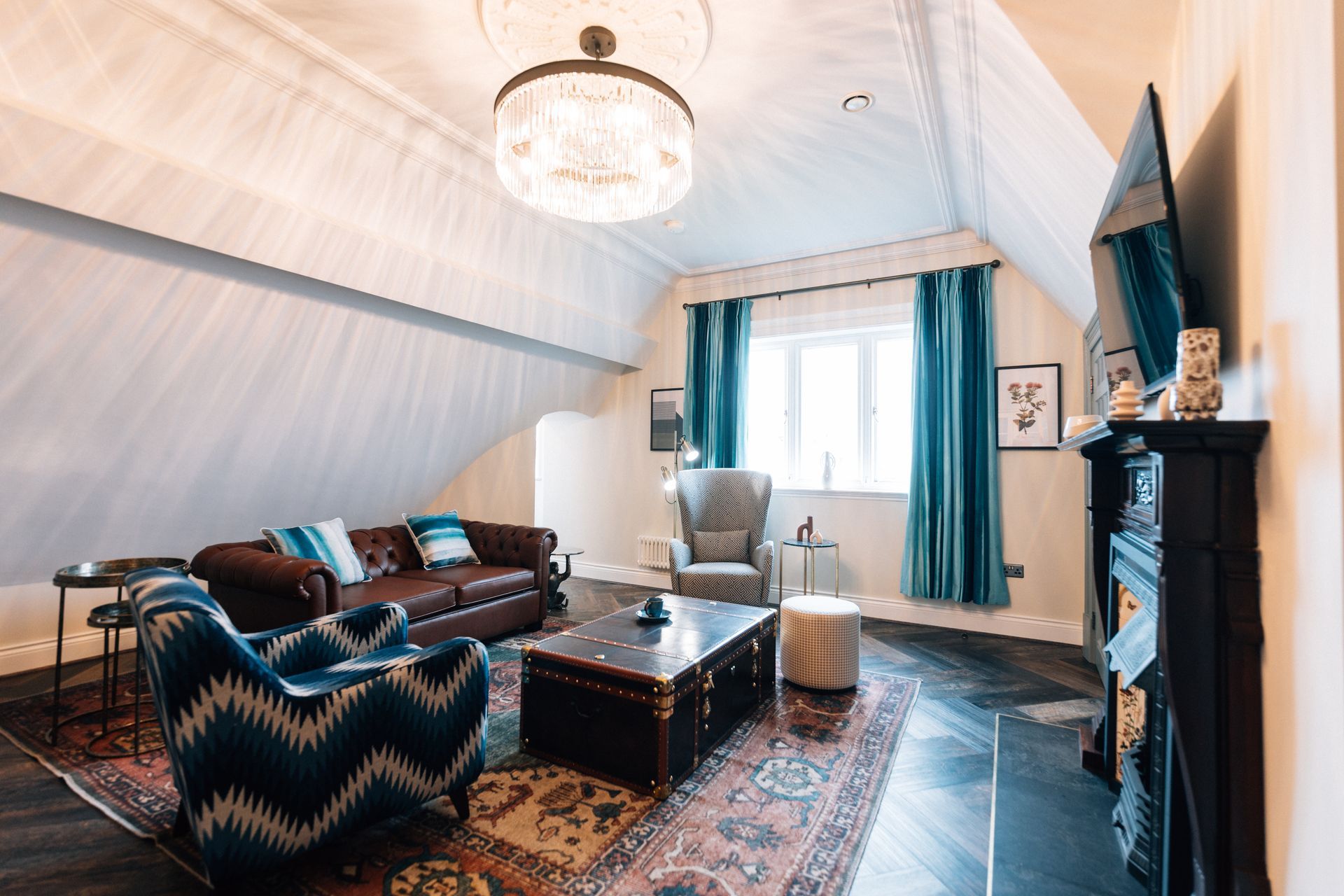 Living room in a slanted-ceiling attic with a brown leather sofa, patterned blue armchair, oriental rug, and chandelier.