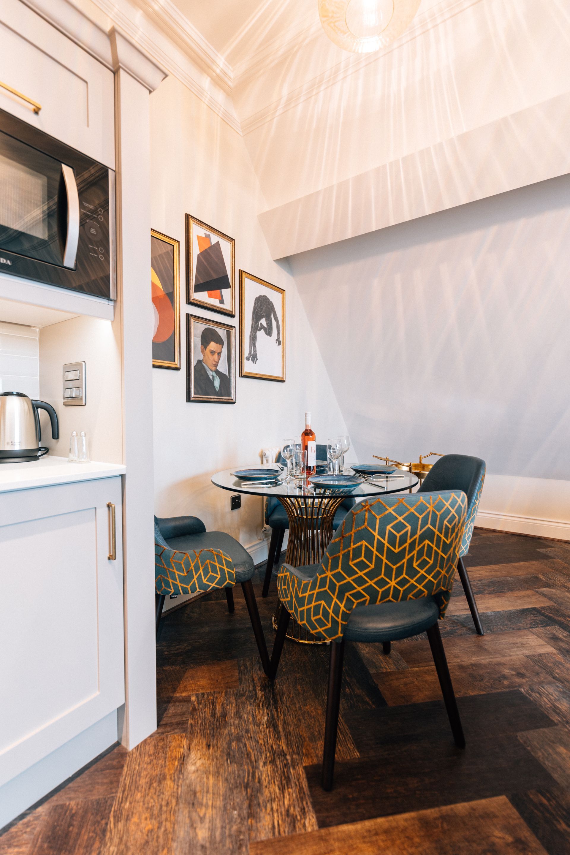 Small dining area with a round glass table, four patterned chairs, and framed portraits on the angled wall. 