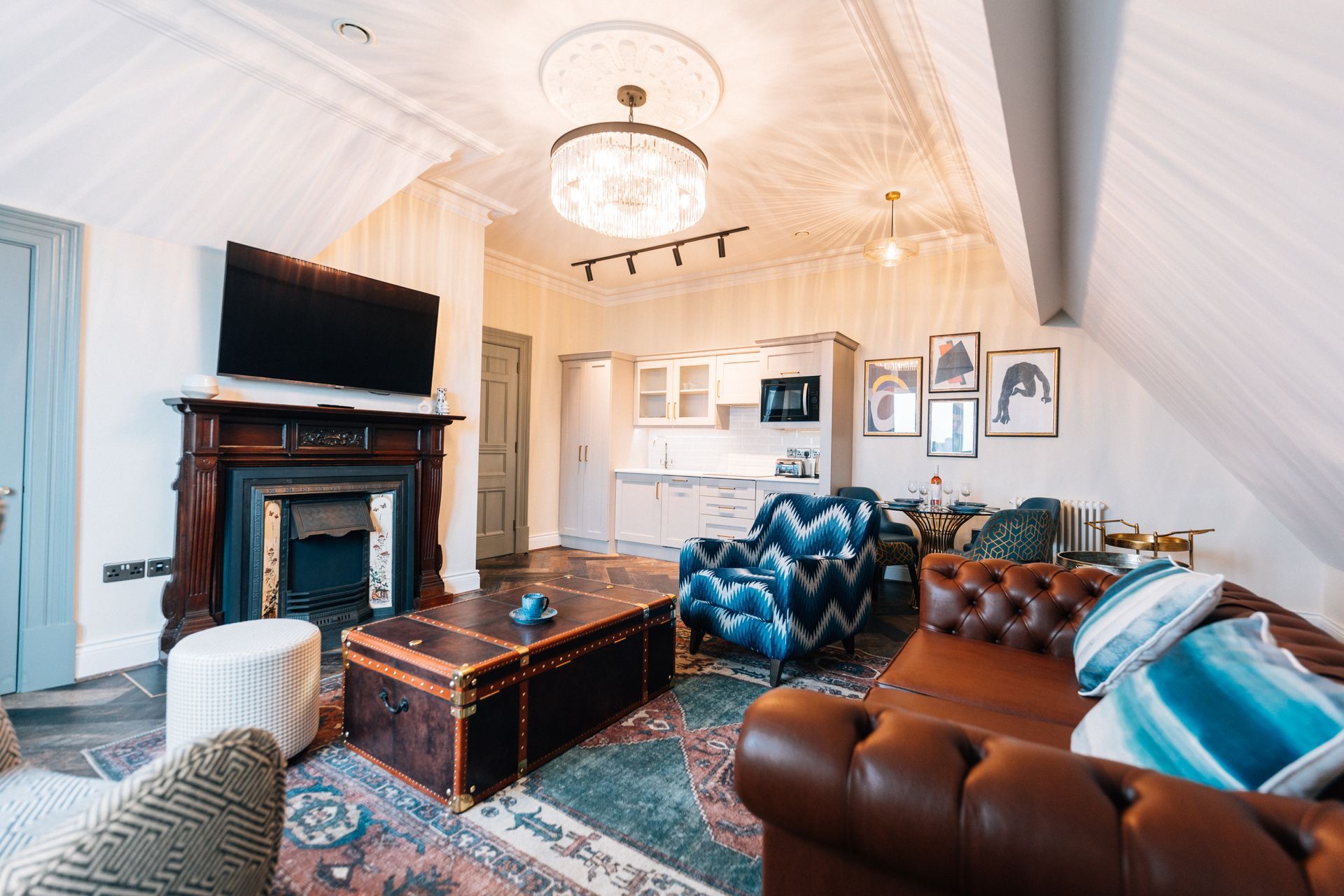 Cozy living room with a fireplace, leather couch, and a trunk coffee table. A blue velvet chair and portraits are visible.