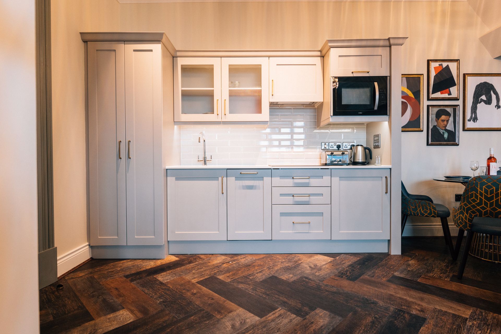 A small, light gray kitchen with white countertops, a microwave, and a herringbone wooden floor. 