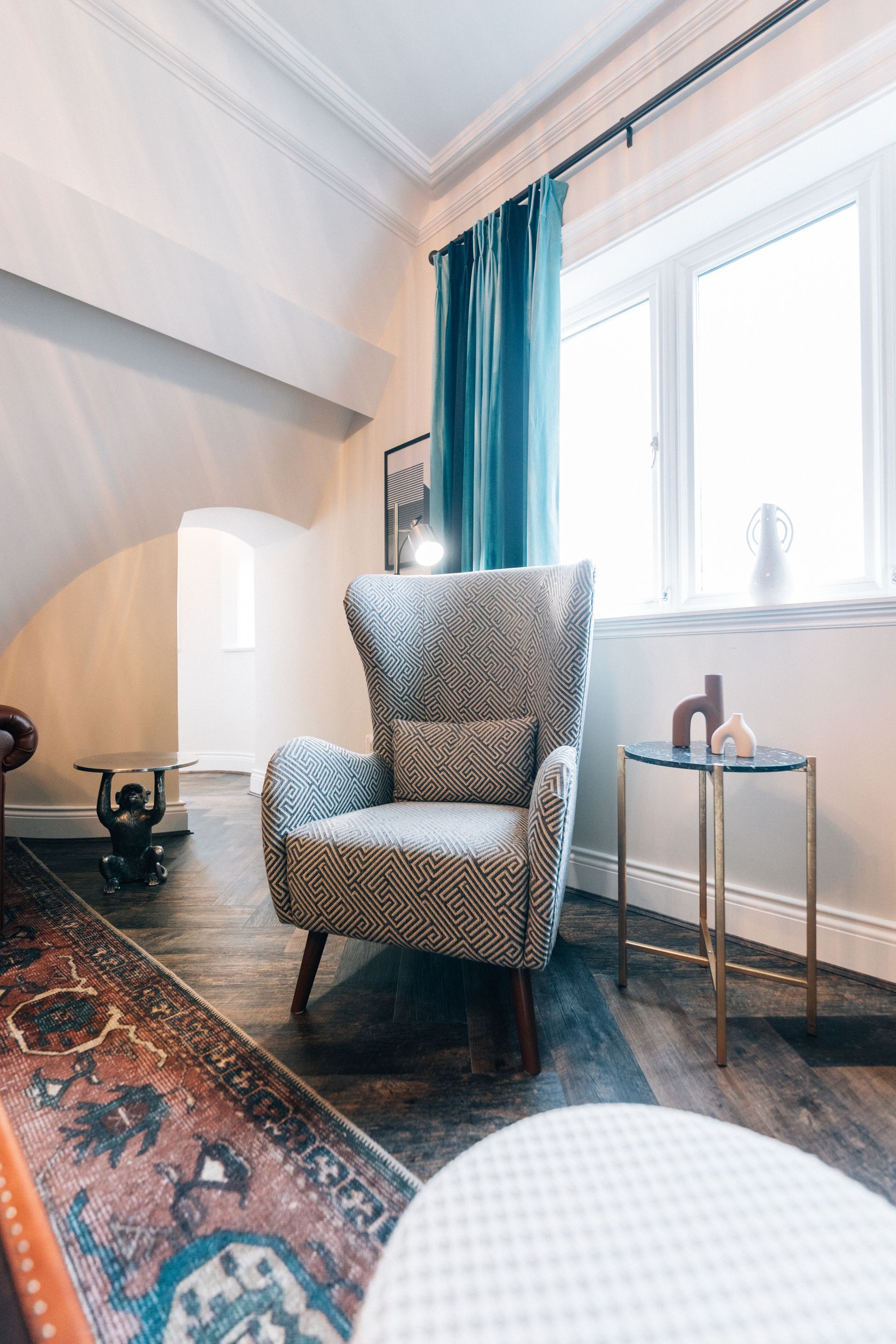 Cozy living room with a patterned armchair, side table, and ornate rug. A window with teal curtains provides natural light.