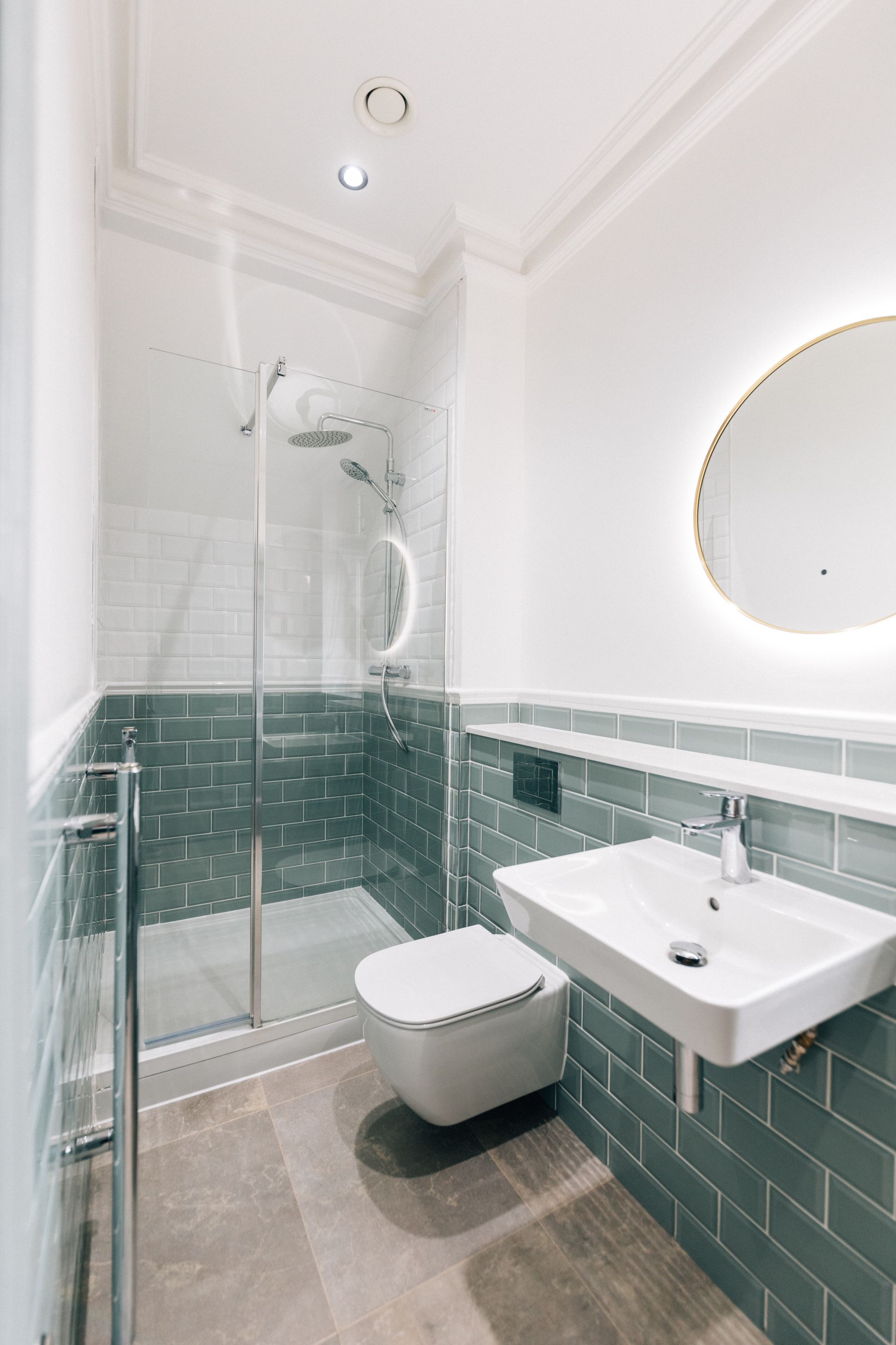 Small modern bathroom with a teal tiled lower wall, white upper wall, shower, toilet, sink, and a round illuminated mirror.
