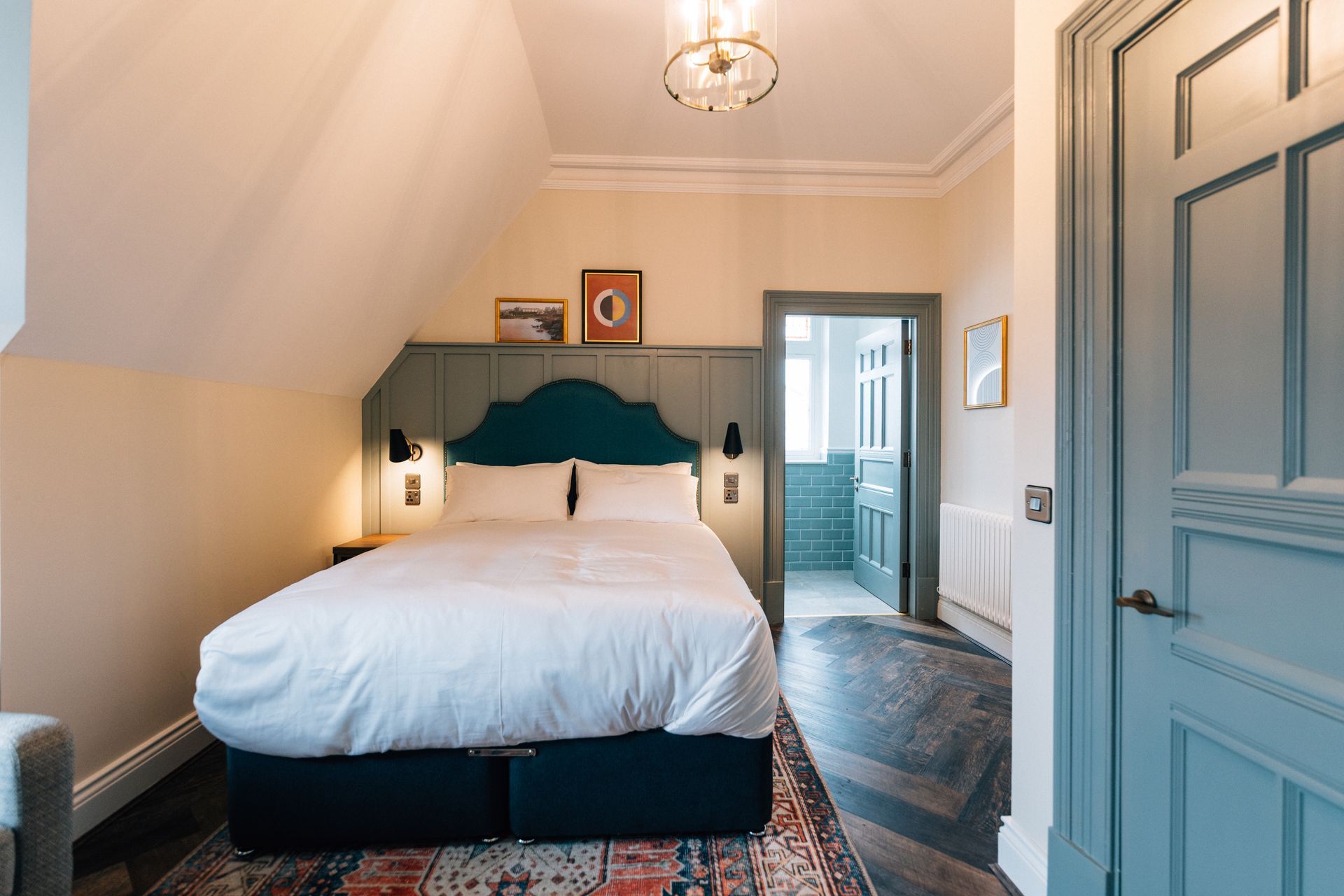 A bedroom with a bed, teal headboard, and doorway to a bathroom. Cream walls, blue accents, and patterned rug.