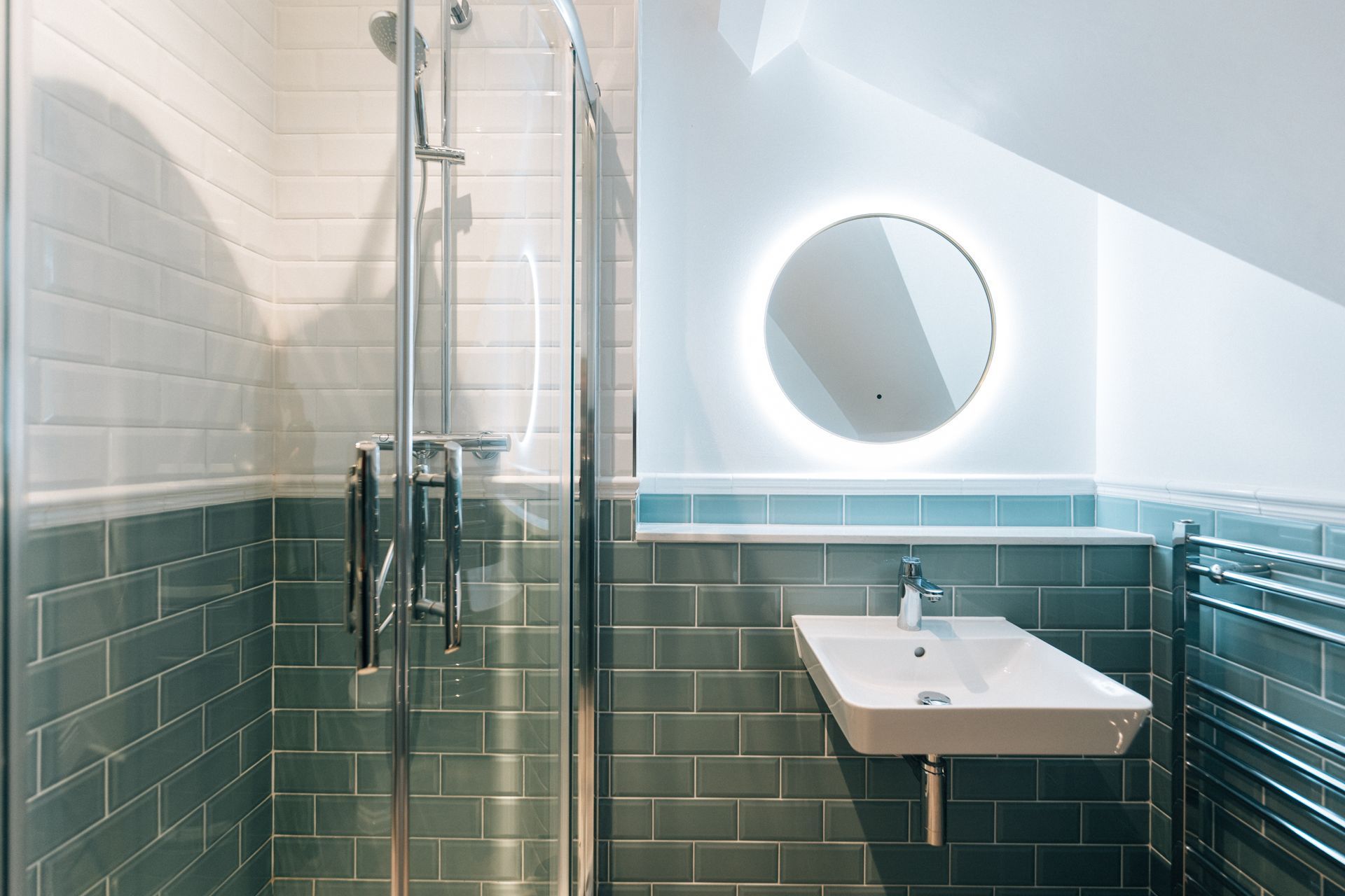 A modern bathroom with blue tiled walls, a glass shower, a floating sink, and a round mirror with a back light.
