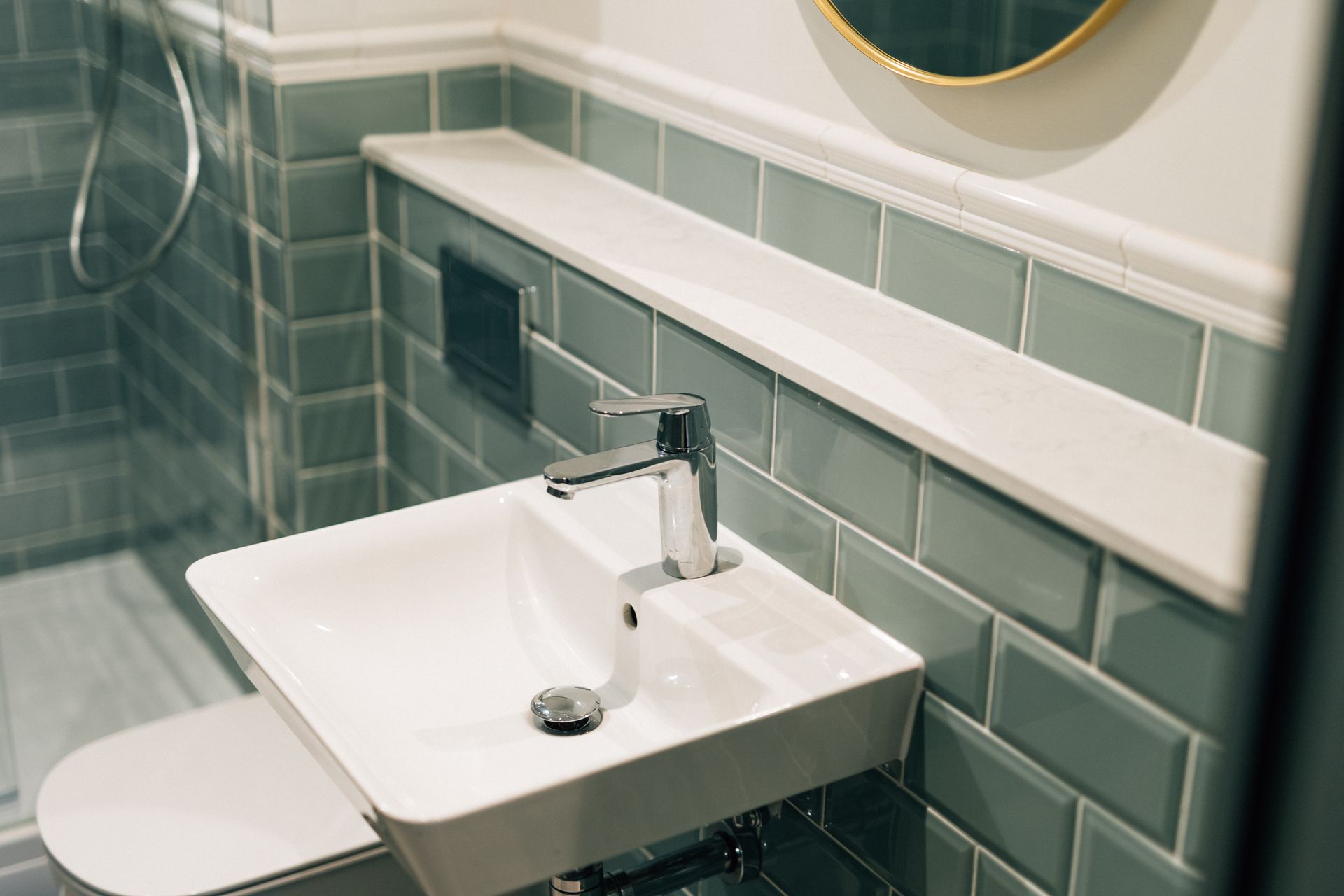 White sink and faucet mounted on a blue tiled wall, with a shelf above. Toilet visible on the left.