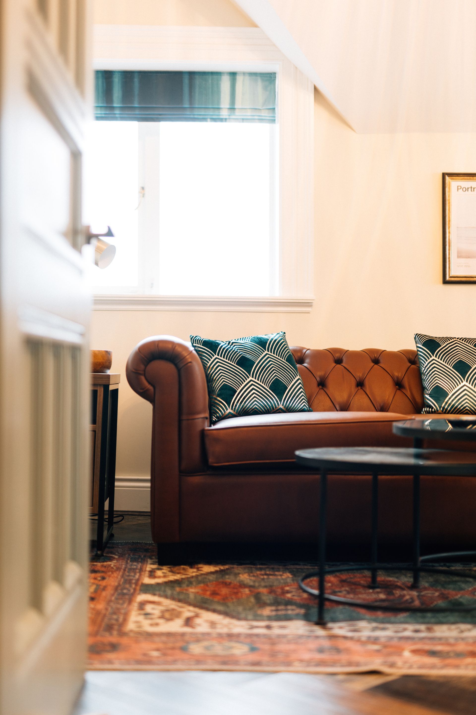A brown leather Chesterfield sofa with teal patterned pillows sits in a room with a rug and a window.