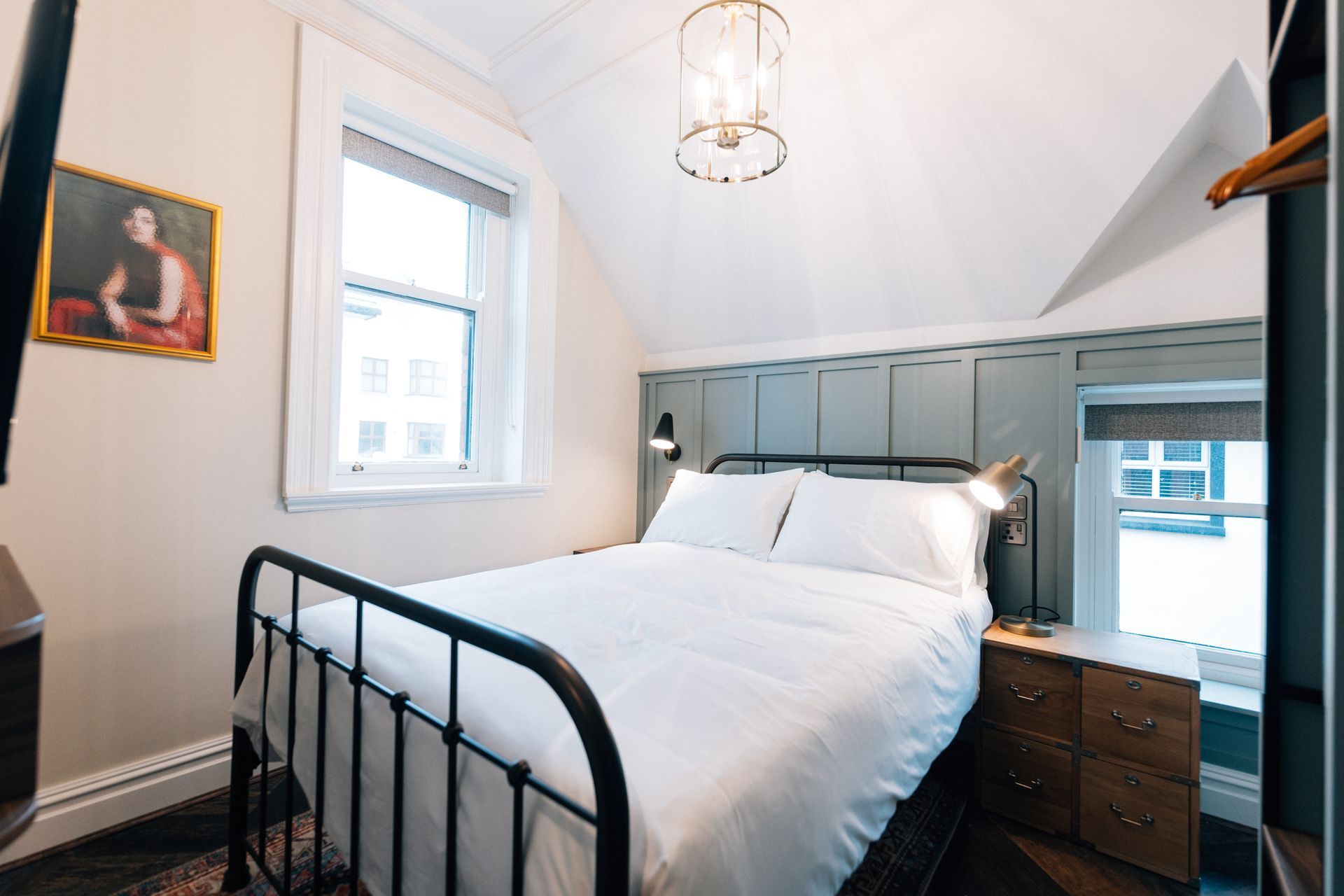 Small bedroom with a bed, window, and painting. Black metal bed frame with white linens. Walls are cream and blue.