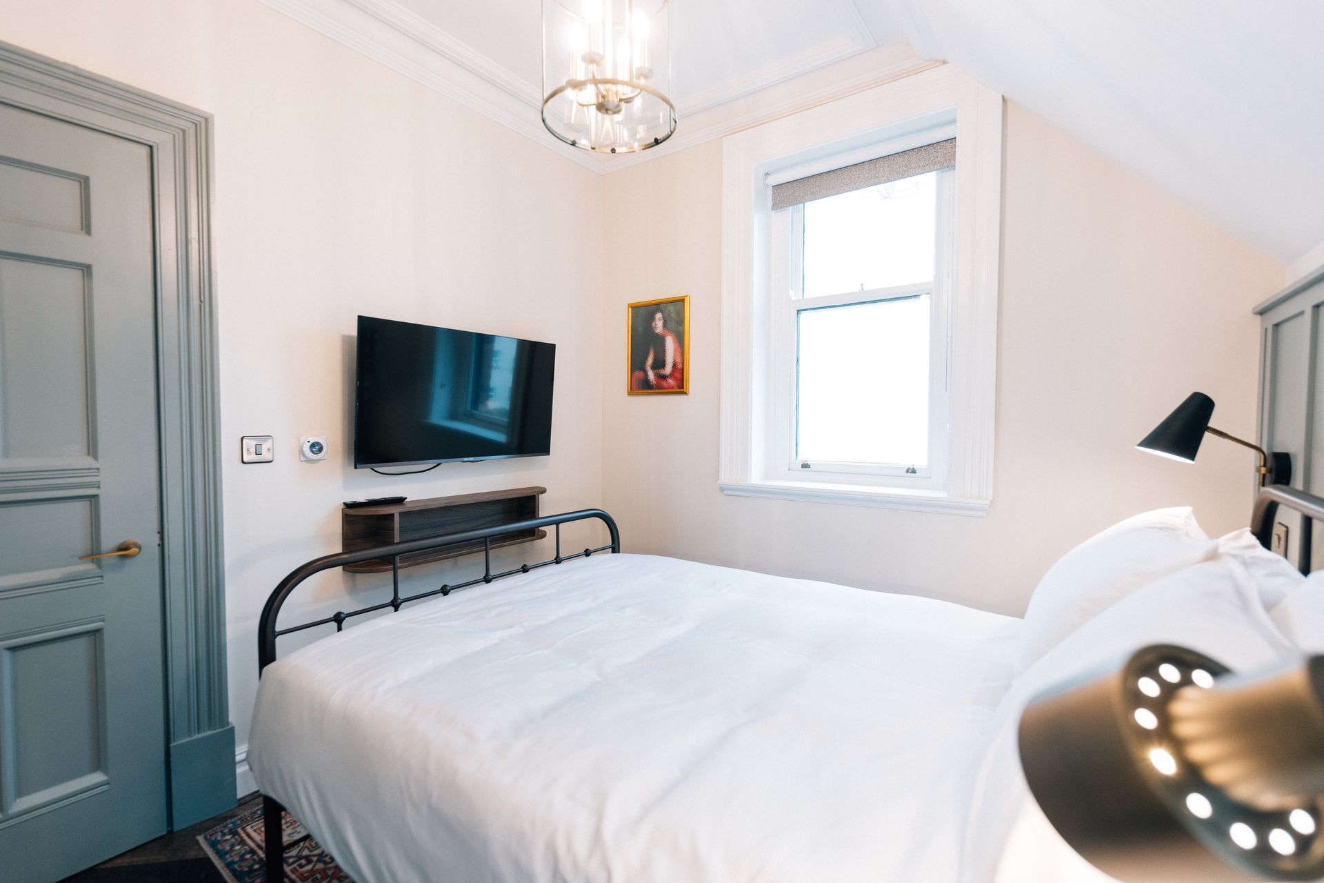 A small bedroom with a bed, TV, and window. The walls are light-colored, and a lamp and a framed picture add detail.