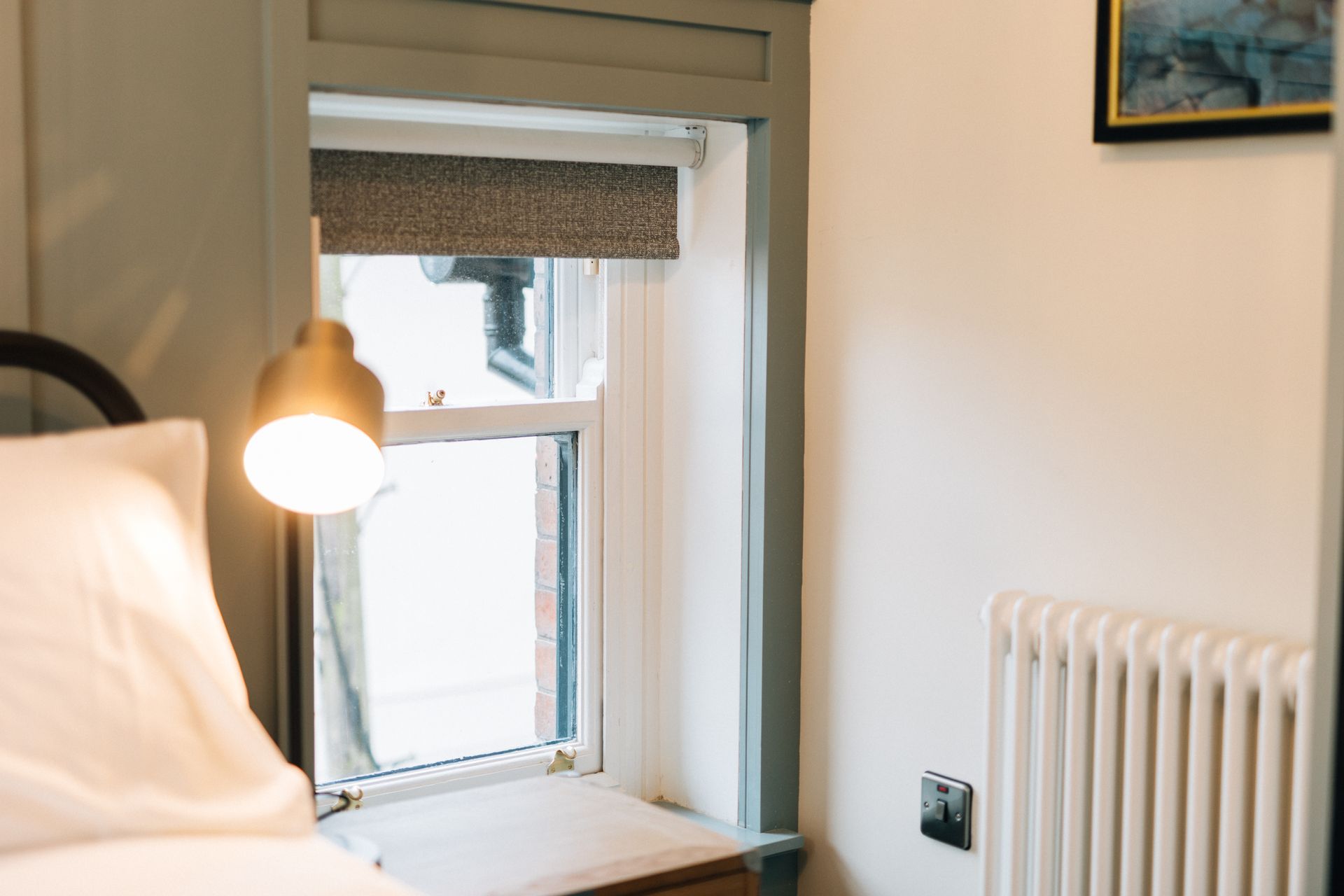 Cozy bedroom corner with a window, radiator, and bedside lamp. Soft light illuminates white bedding and wall.