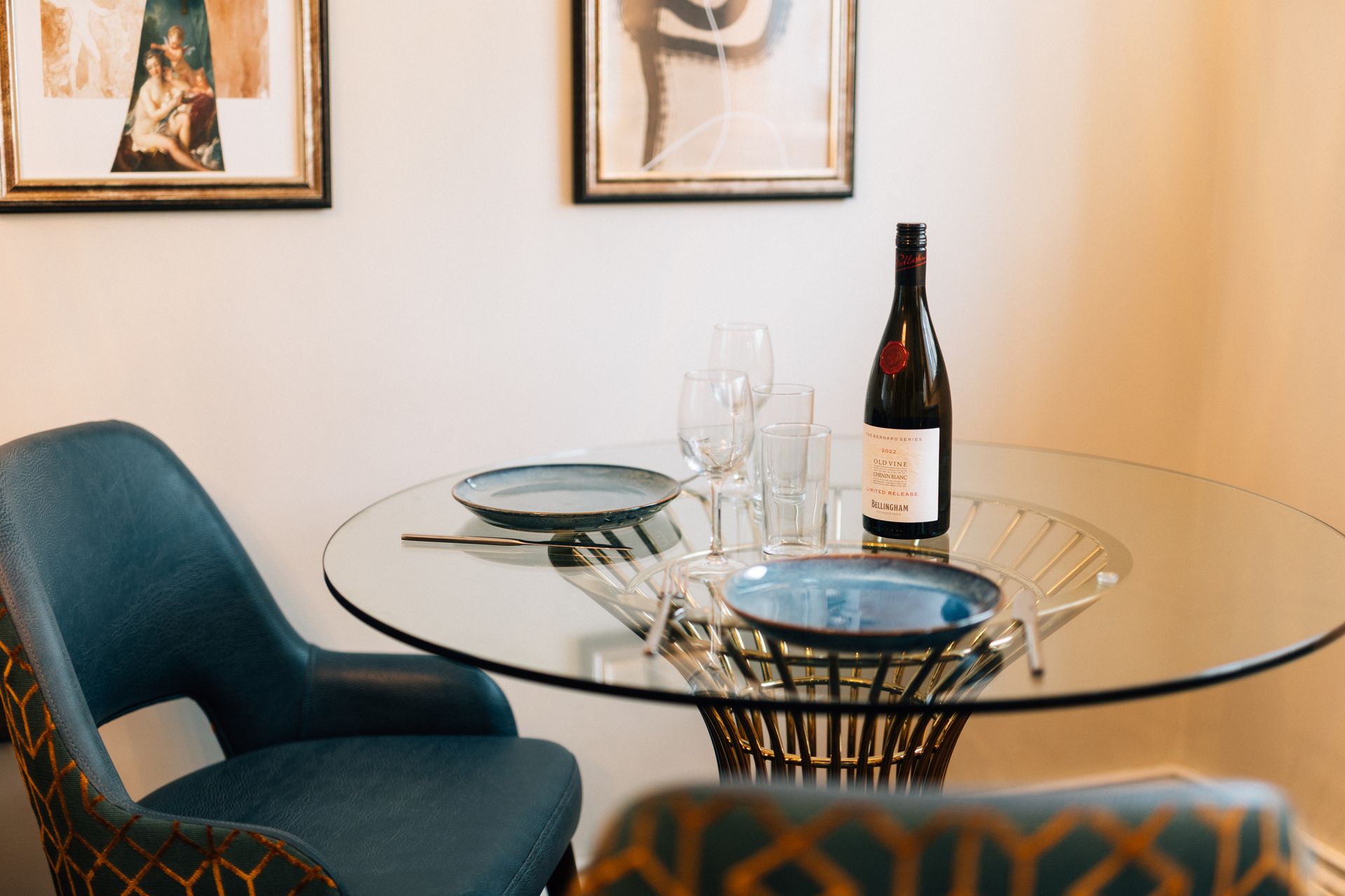 A dining table set for two with blue plates, wine glasses, and a bottle of red wine; two blue chairs.