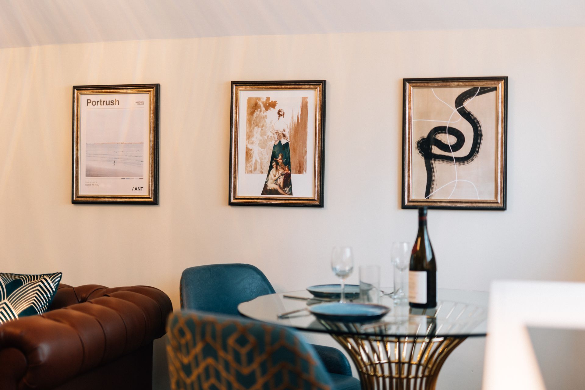 Dining area with three framed artworks over a glass table set for two. A brown leather sofa is visible.