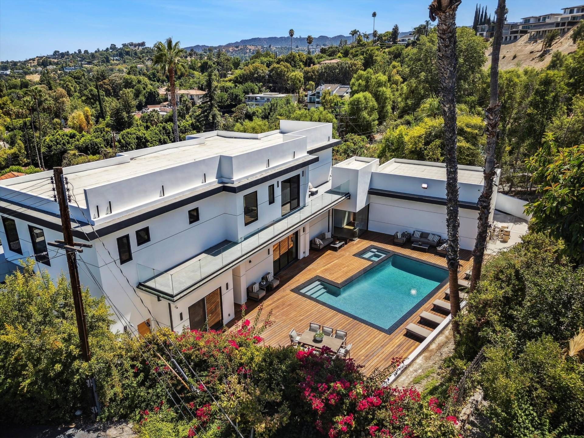 Modern white house with a pool, wooden deck, and lush greenery in a hillside setting.
