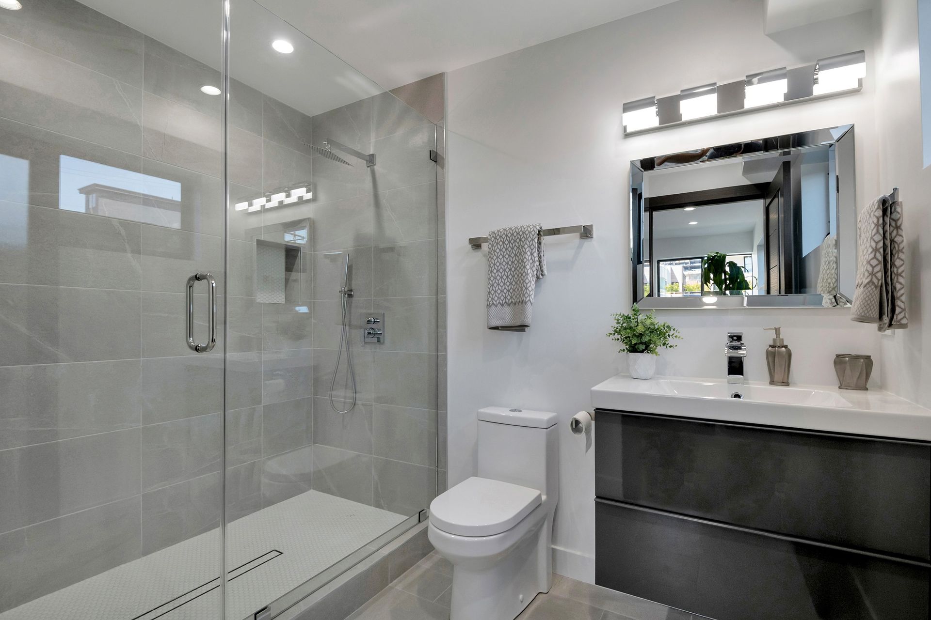 Modern bathroom with gray tiled shower, toilet, and vanity. White walls and chrome fixtures.