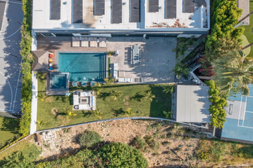 Aerial view of a modern house with a pool, patio, and tennis court, surrounded by greenery.