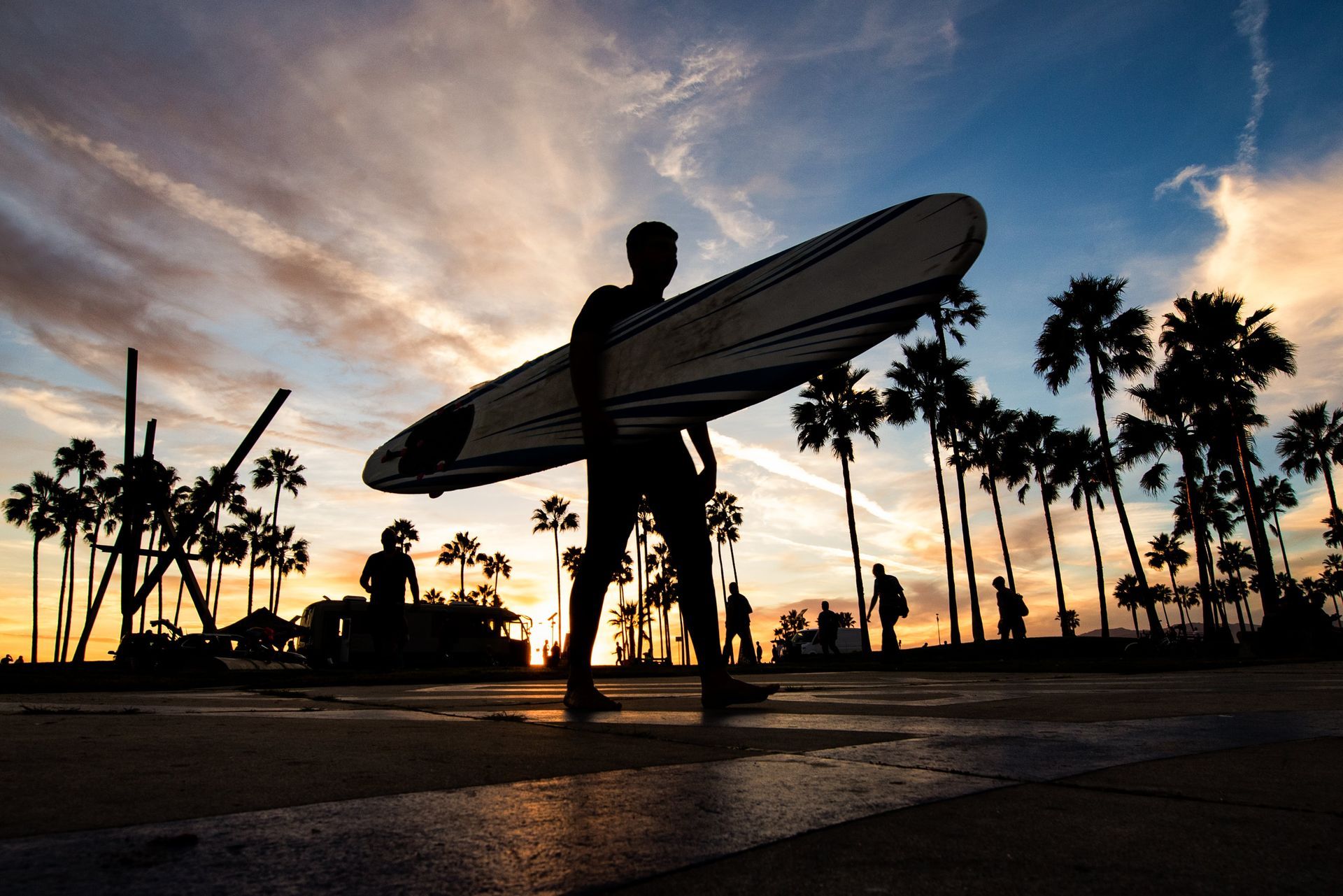 A man is carrying a surfboard on a sidewalk at sunset.